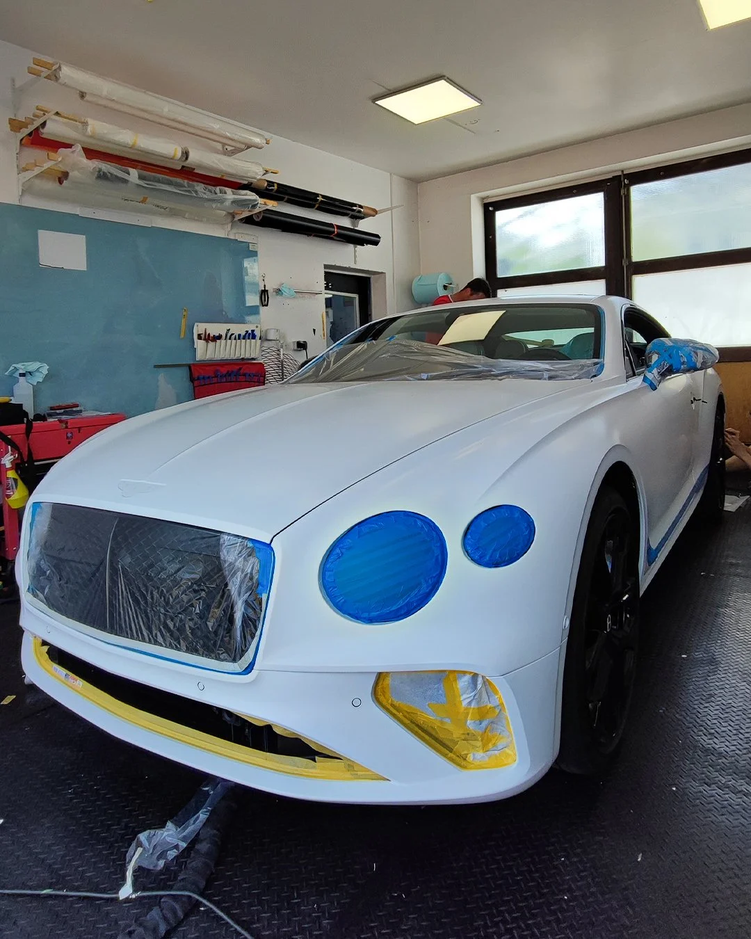 Ein weißer Sportwagen, der für den Lackierprozess vorbereitet wird, mit blauer und gelber Abdeckung auf den Scheinwerfern und anderen Teilen, in einer Werkstatt.