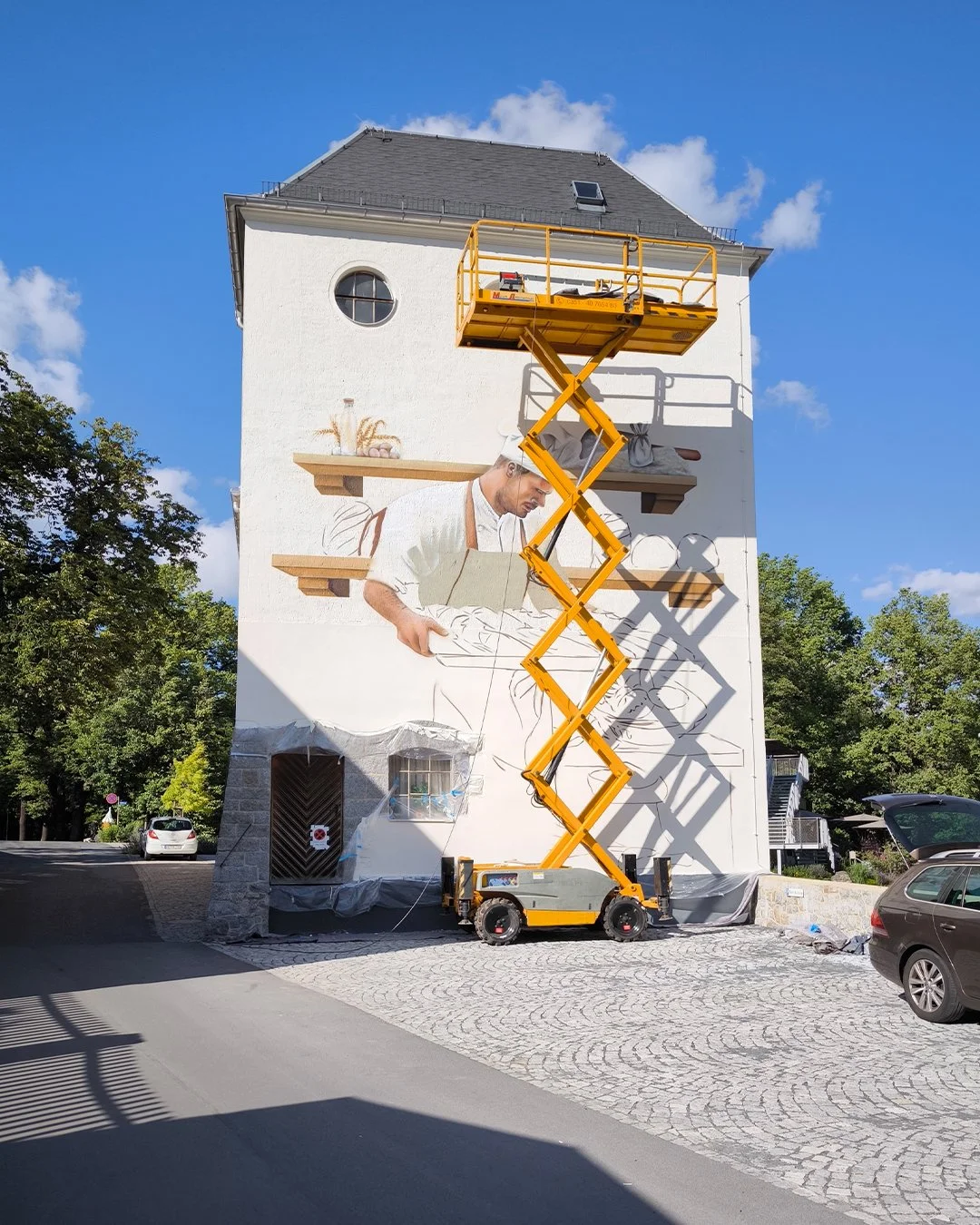 Fassade eines Gebäudes, das eine große Wandmalerei eines Kochs beim Kochen zeigt, mit einer Hebebühne davor, die die Malerarbeiten ermöglicht.