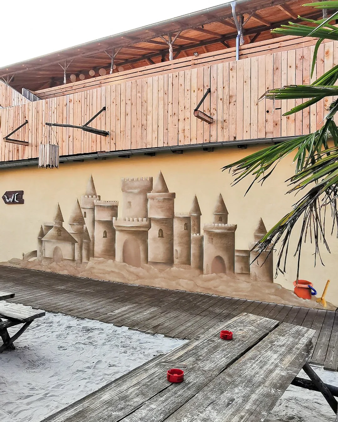 Sandtura im Kinderbereich mit Burgmauer auf einem Sandboden, umgeben von Holzterrasse, mit Sandspielzeug und Palmen.
