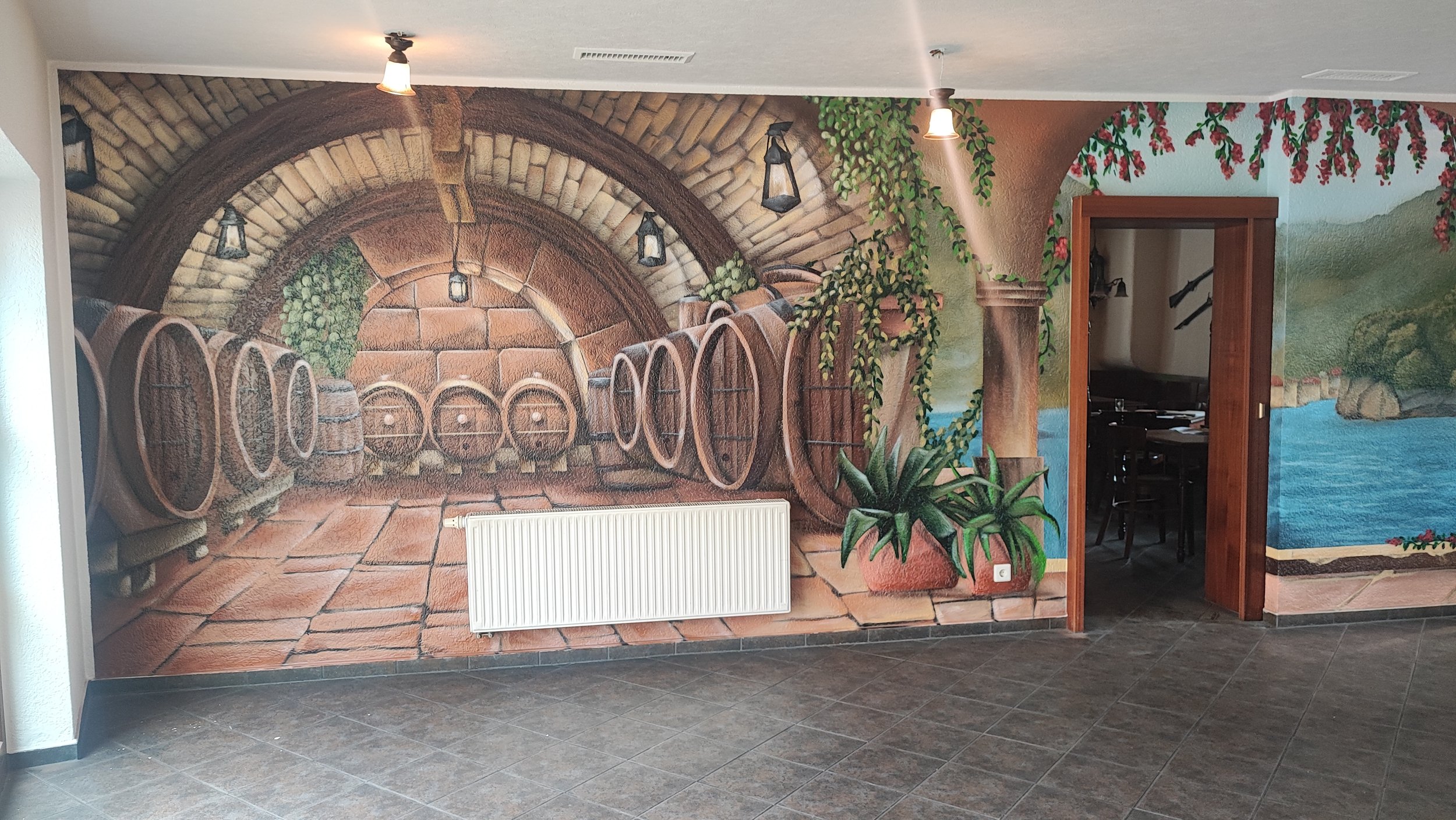 Wandbild mit einer Kellerkulisse mit Holzbottichen, Pflanzen und einer Steinmauer, daneben offenes Zimmer mit einem Tisch und Stühlen, Wandmalerei eines Sees und grüner Hügellandschaft.