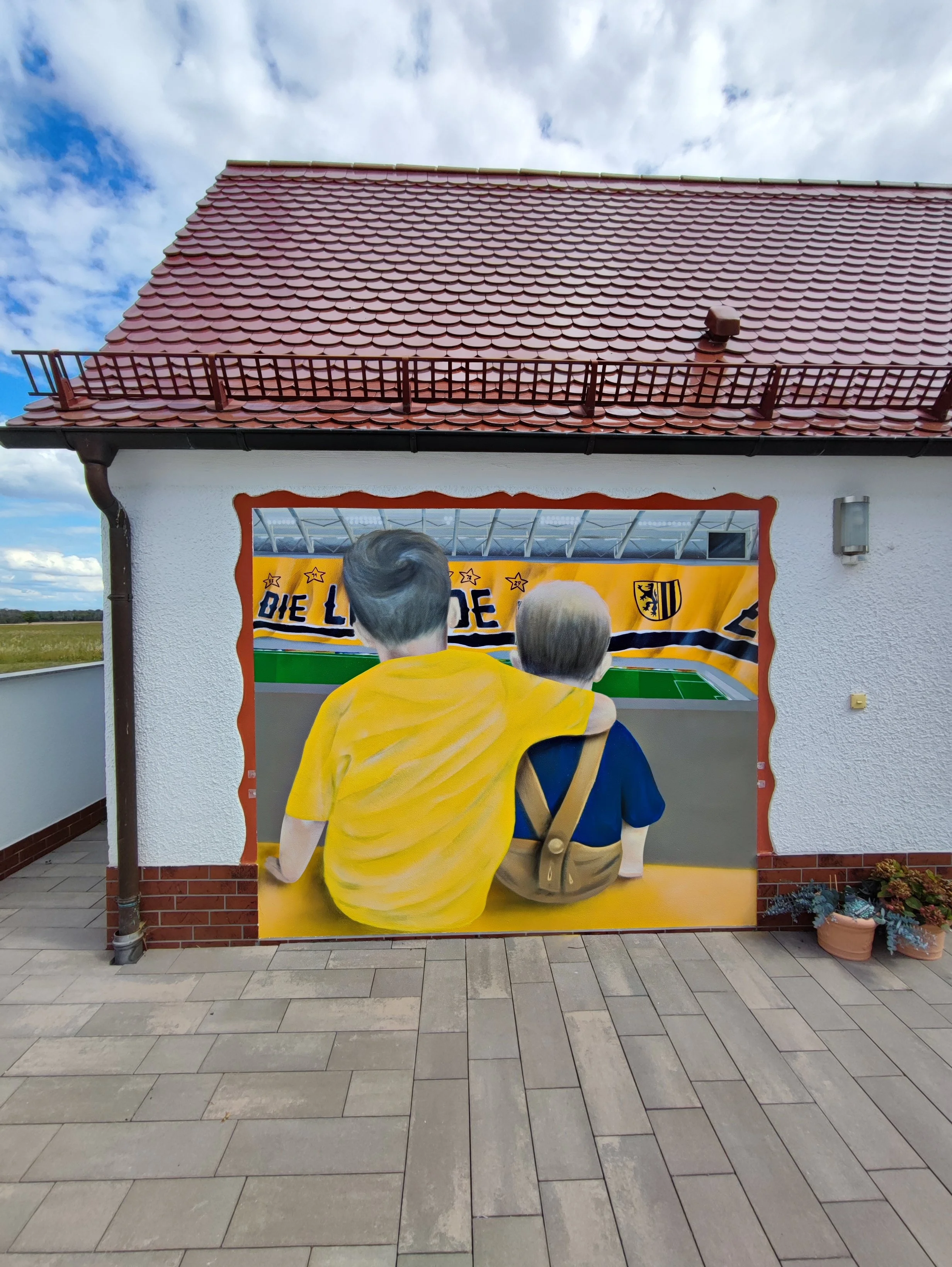 Fassade eines Hauses mit Wandgemälde eines Mann und einem Jungen, die zusammen auf ein Fußballspiel schauen, vor einem gelben Banner mit deutschem Fußballverein.