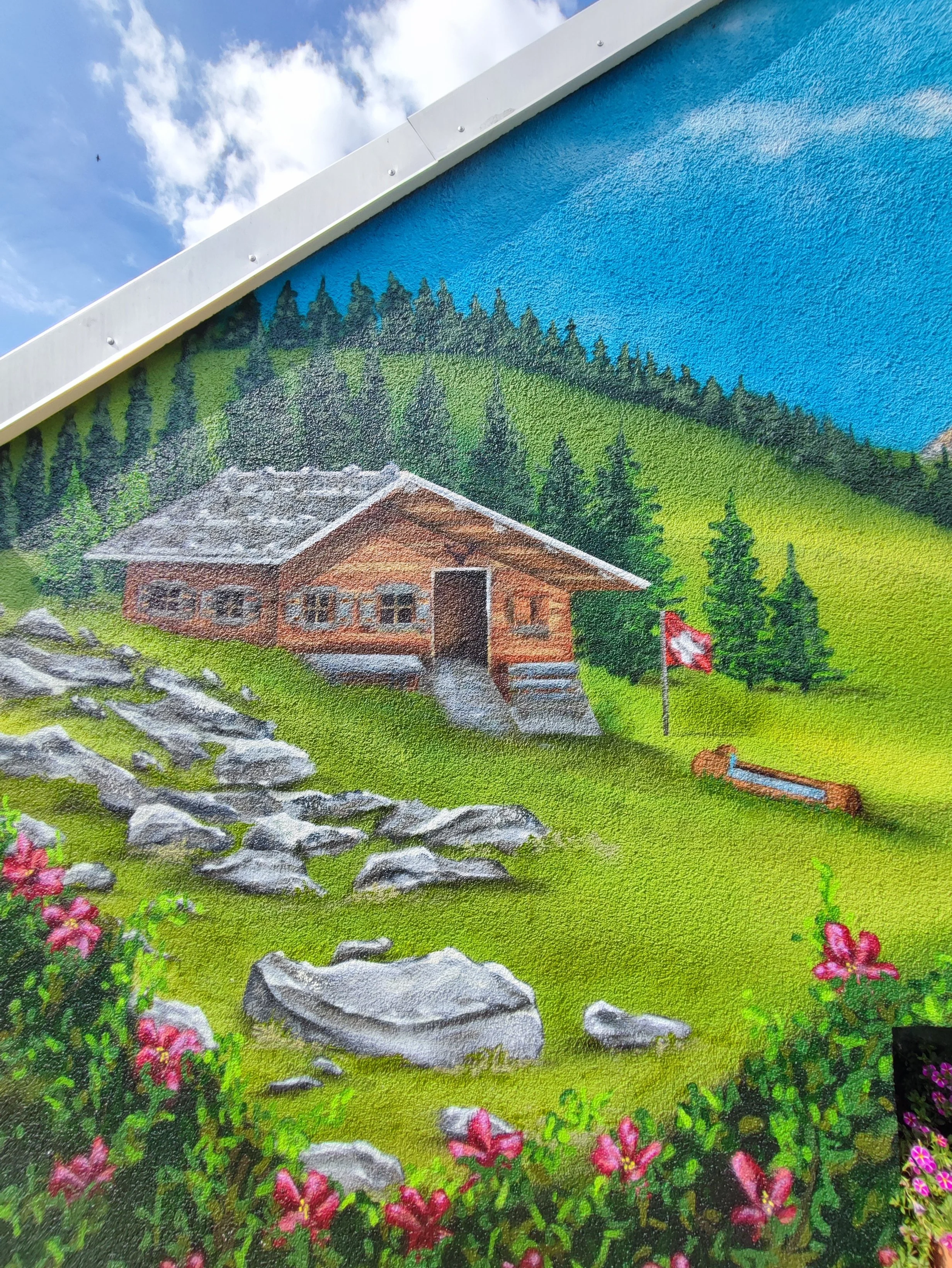 Eine bunte Wandmalerei zeigt eine Berghütte auf grüner Wiese mit bunten Blumen, einem Schweizer Fähnchen, einem Wasserbehälter und einer Felslandschaft, im Hintergrund ein Baumhügel und ein blauer Himmel mit Wolken.