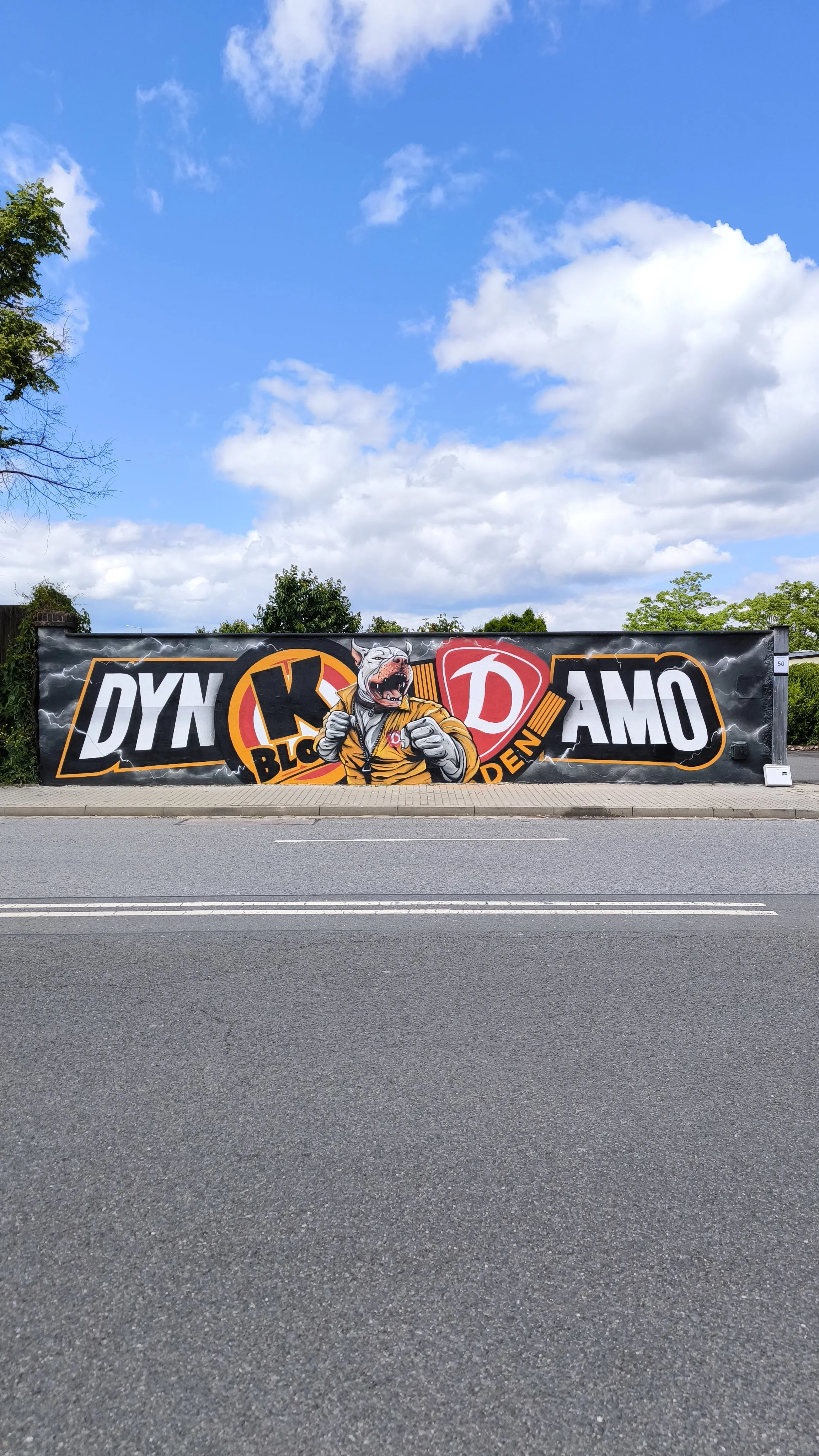 Werbetafel mit einem kämpfenden Bulldoggen-Charakter, der eine Faust zeigt, und den Worten 'DYN', 'K', 'D', und 'AMO' auf einem schwarzen Hintergrund mit einem blauen Himmel im Hintergrund.