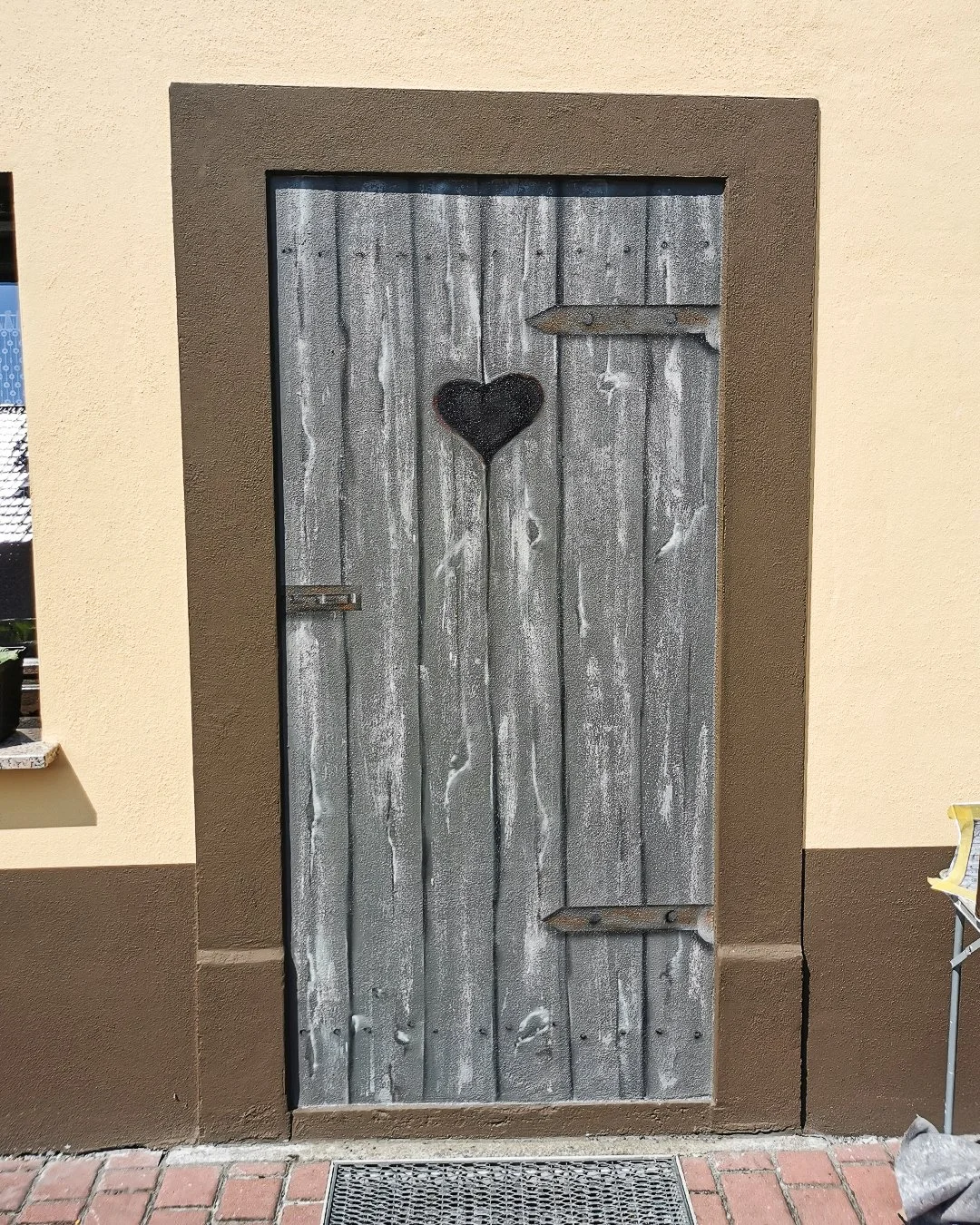 Eine alte Holztür mit einem schwarzen Herz in der Mitte. Die Tür befindet sich in einem braunen Rahmen vor einer cremefarbenen Wand.