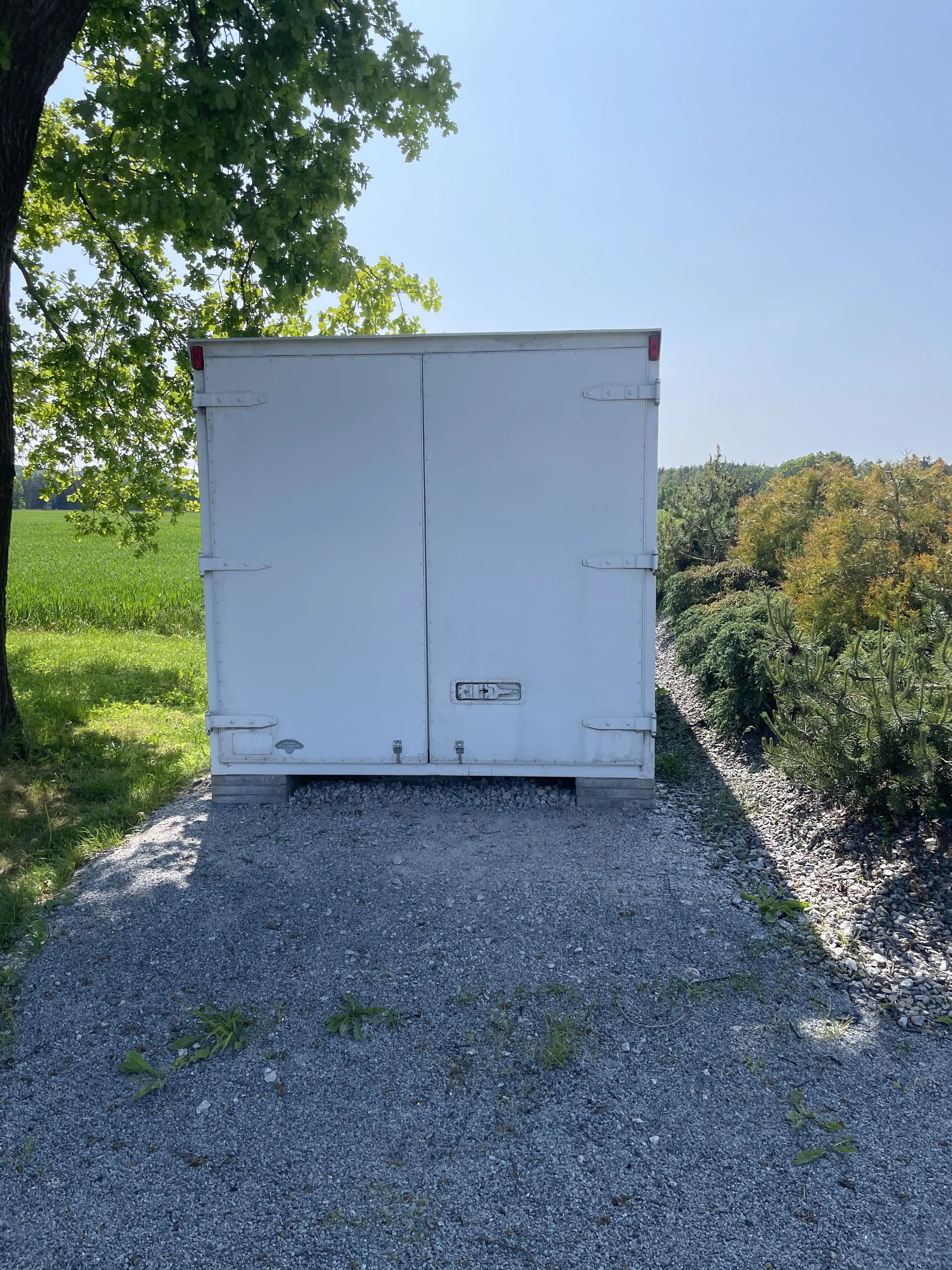 Ein weißer Lastwagen mit geschlossenen Türen steht auf einer Kiesstraße, flankiert von Bäumen und Feldern bei schönem Wetter.