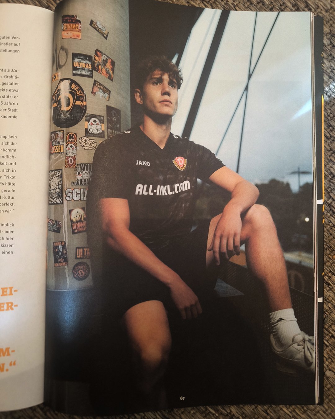 Ein junger Mann in einem schwarzen Sporttrikot sitzt auf einer Bank, im Hintergrund eine Wand mit Stickern und eine Glasscheibe, bei Sonnenuntergang im Freien.