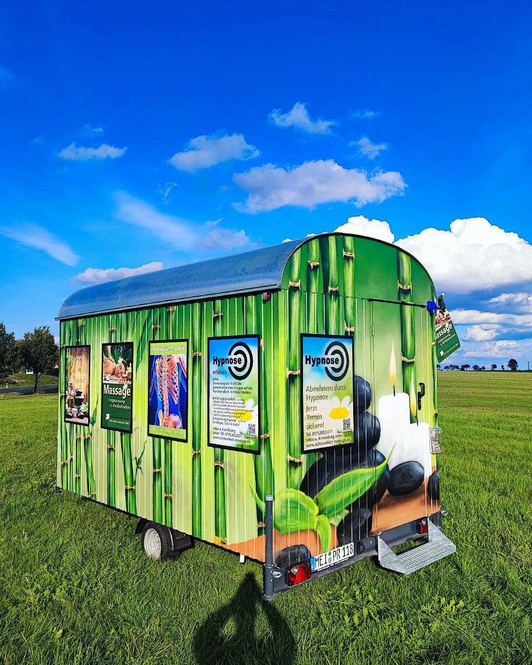 Mobiles Werbe- und Informationsschild mit Bildern von Massage, Hypnose und Natur, auf einem grünen Feld mit blauem Himmel und Wolken im Hintergrund.