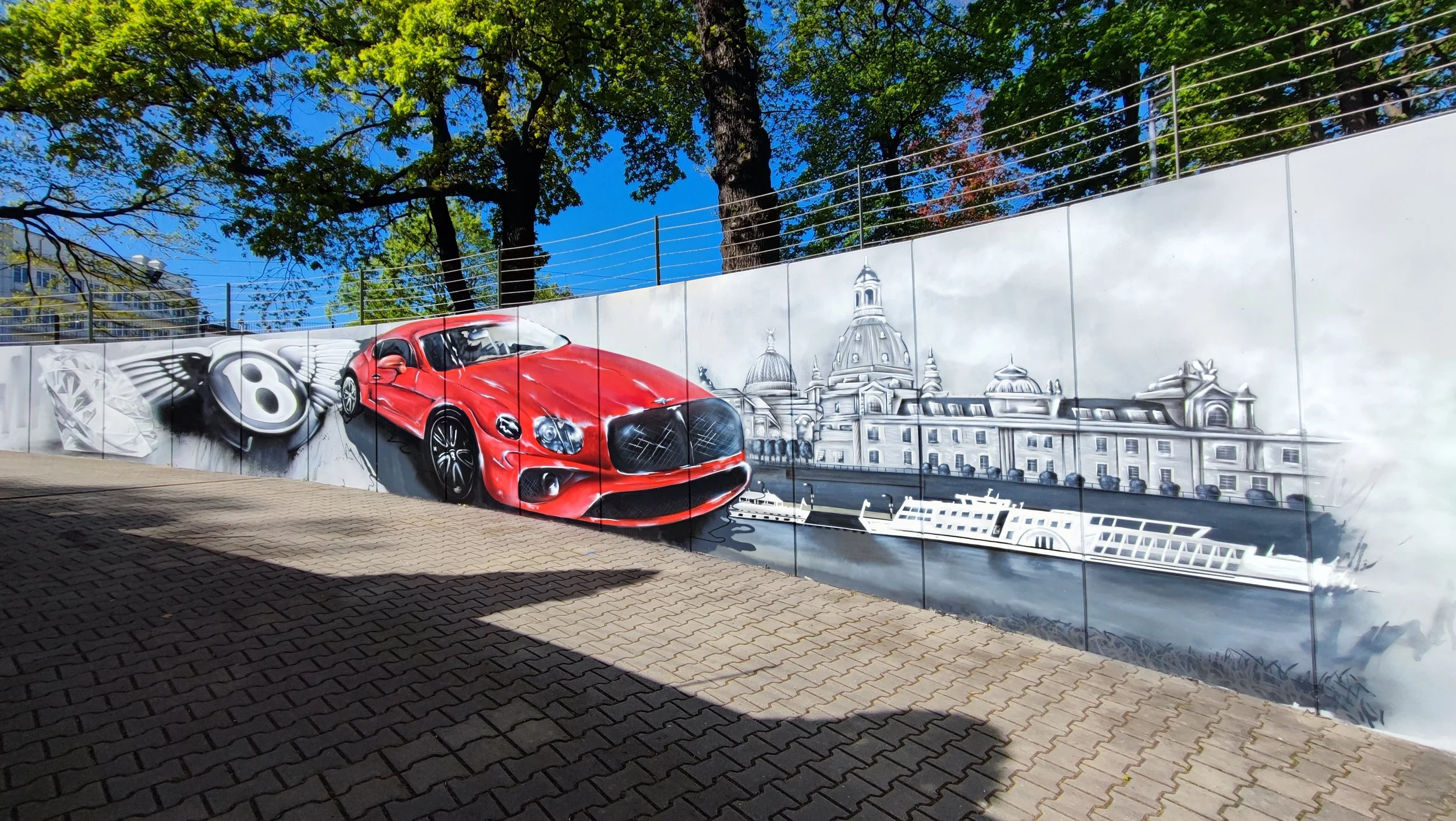 Wandmalerei mit rotem Sportwagen, Stadtansicht, Boot und Skyline im Hintergrund, in Grautönen, bei Sonne und Schatten auf gepflastertem Boden.