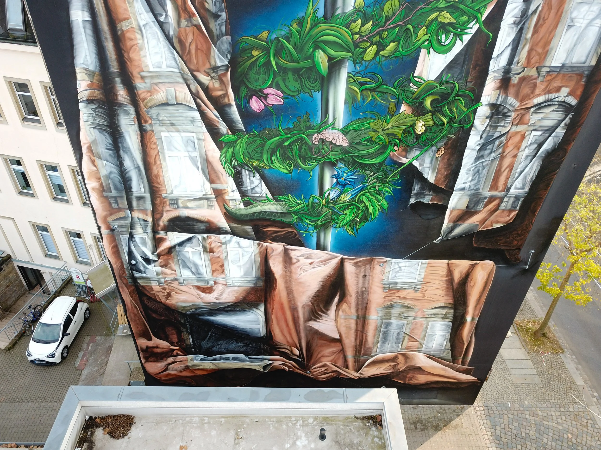 Buntes Wandbild an der Seite eines Gebäudes, das eine große grüne Pflanze mit pinker Blüte und blauen Vogel zeigt, umgeben von naturähnlichen Elementen und einem Himmel mit blauen Hintergrund.