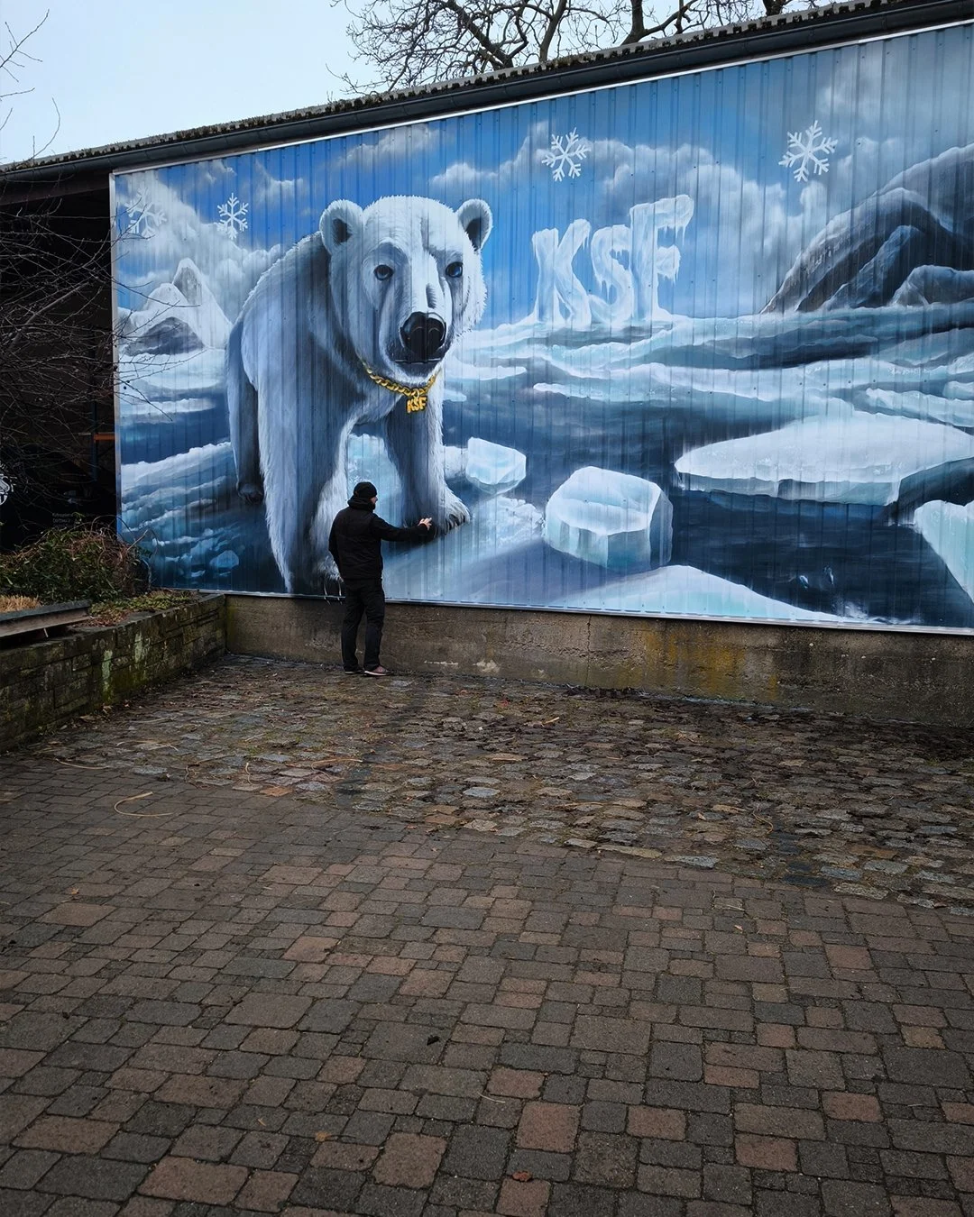 Ein Mann steht vor einem Wandgemälde eines Eisbären in einer arktischen Landschaft mit Eis und Schnee. Das Gemälde zeigt den Eisbären groß und realistisch dargestellt, mit der Beschriftung 'KSE' im Hintergrund.