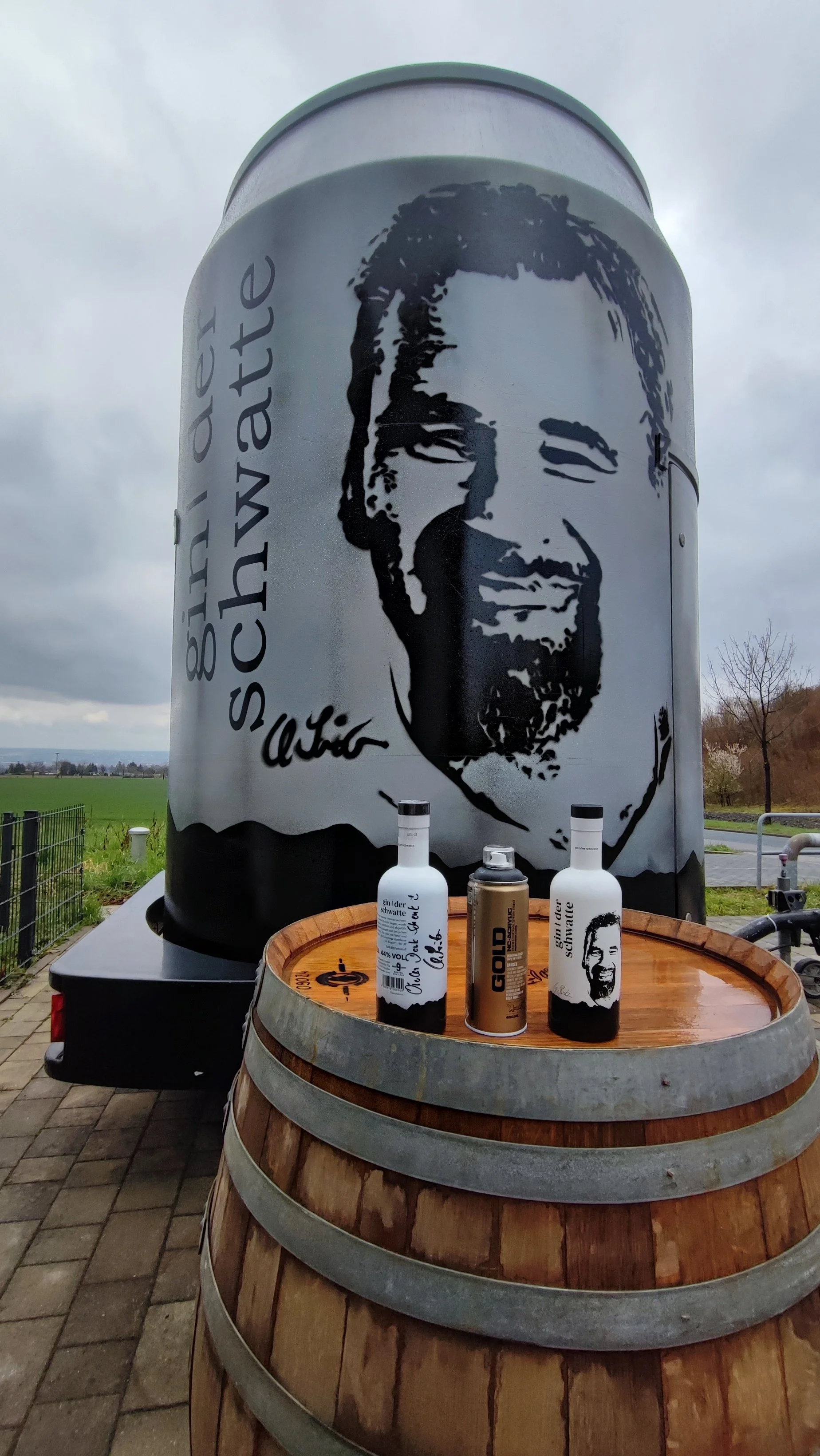Großer Metalltank mit Schwarzweiß-Porträt eines lachenden Mannes, daneben Text in deutscher Sprache und Signatur, auf einer Weinholzrolle stehen zwei Flaschen und eine Dose, im Freien mit bewölktem Himmel und grüner Wiese im Hintergrund.