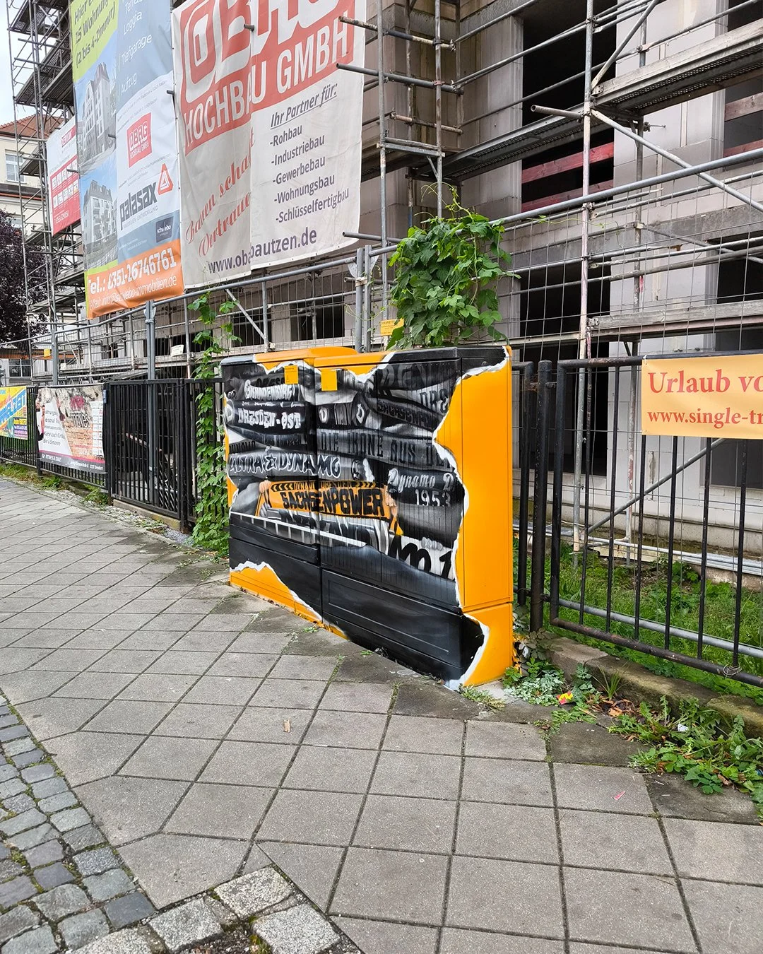Straßenansicht mit einem Zaun, Baugerüsten und mehreren Werbetafeln, sowie einem abgestellten, bemalten Kasten, der Graffiti und Werbung trägt.