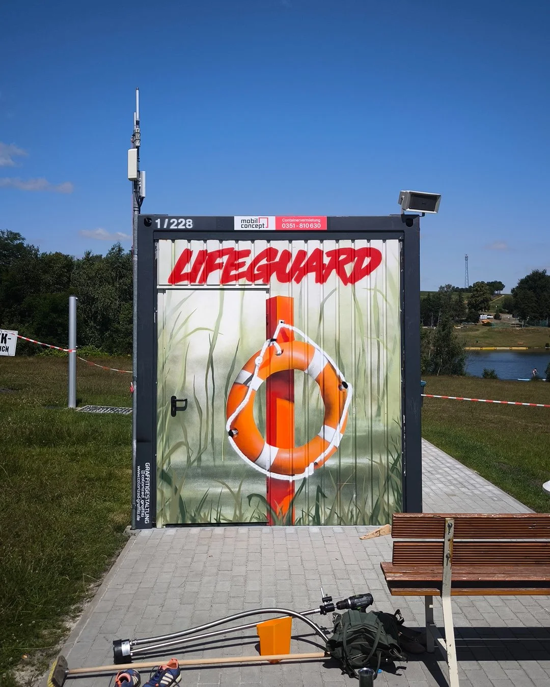 Ein Rettungsschirm mit dem Wort ‚Lifeguard‘ in roter Schrift, vor einer Boje, mit Wasser im Hintergrund, bei sonnigem Himmel.