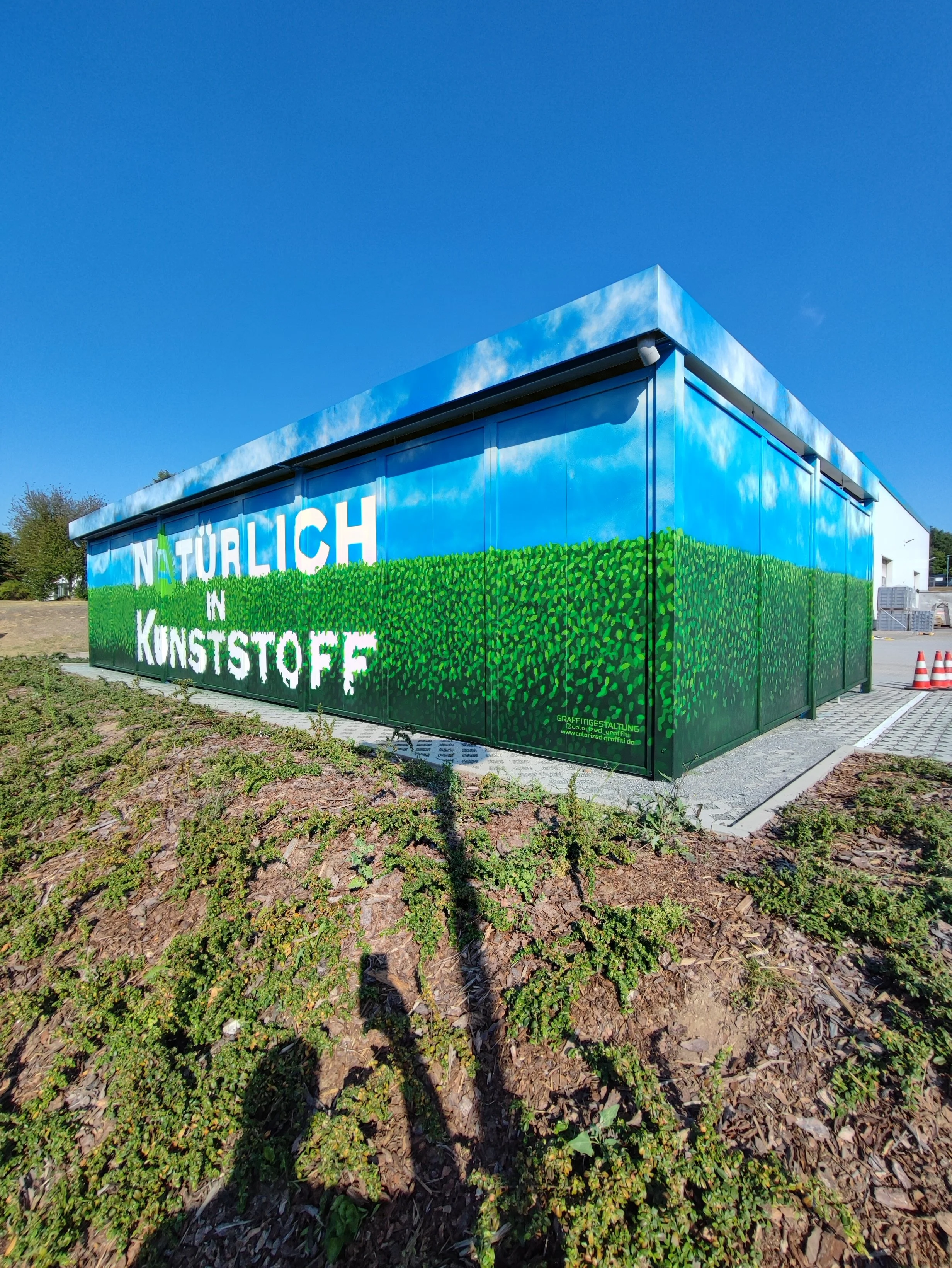 Gebäude mit Wandmalerei, die den Himmel und eine grüne Hecke zeigt, mit dem Text 'Natürlich in Kunststoff'