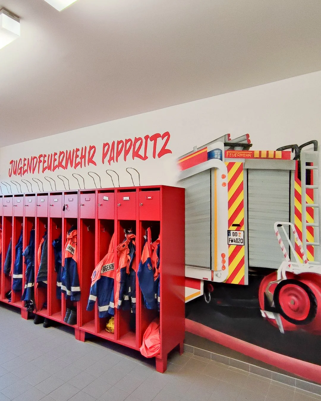 Feuerwehrjacken und -helme in rote Spinde in einer Feuerwehrhalle, mit der Aufschrift „Jugendfeuerwehr Papritz“ an der Wand, daneben ein gemaltes Feuerwehrfahrzeug.