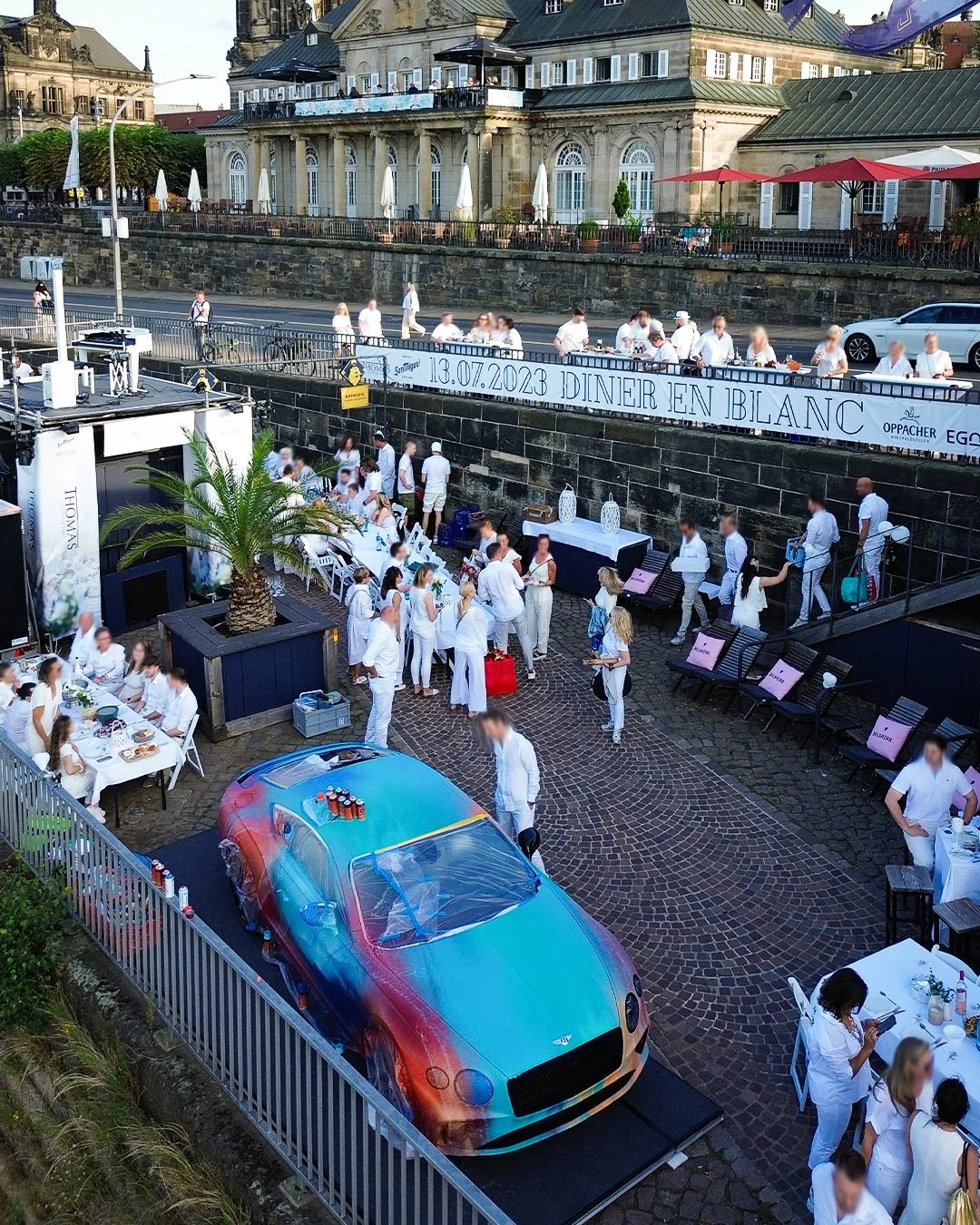 Ein Sideways-Luftaufnahme zeigt einen gehobenen Gartenbereich mit vielen Menschen in weißen Kleidern, die bei einem Event oder einer Feier zusammen sitzen und essen. Es gibt einen limitierten, bunt bemalten Sportwagen, der am Boden montiert ist, sowi