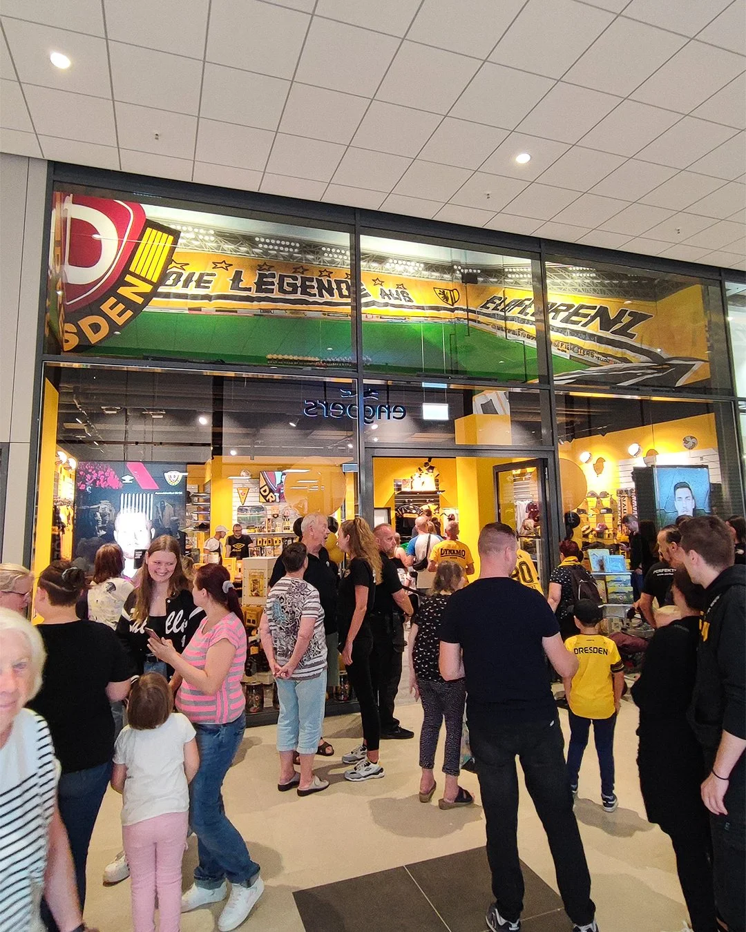 Menschen warten in einer Rabatt- und Fanartikel-Geschäft mit gelber Dekoration, spezialisiert auf den Fußballclub Dynamo Dresden. https://www.dynamo-fanshop.de/
