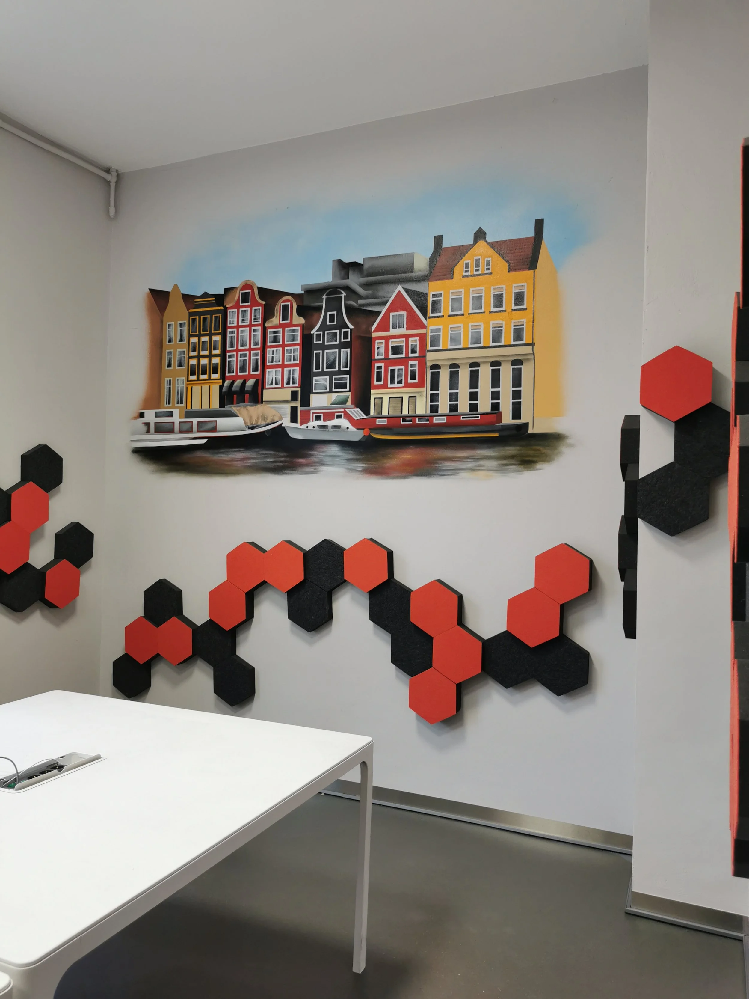 Wand mit Wandkunst von bunt gestrichenen Häusern und Wasserhäusern, dekorative schwarze und rote sechseckige Akzente an der Wand, weißer Tisch im Vordergrund.