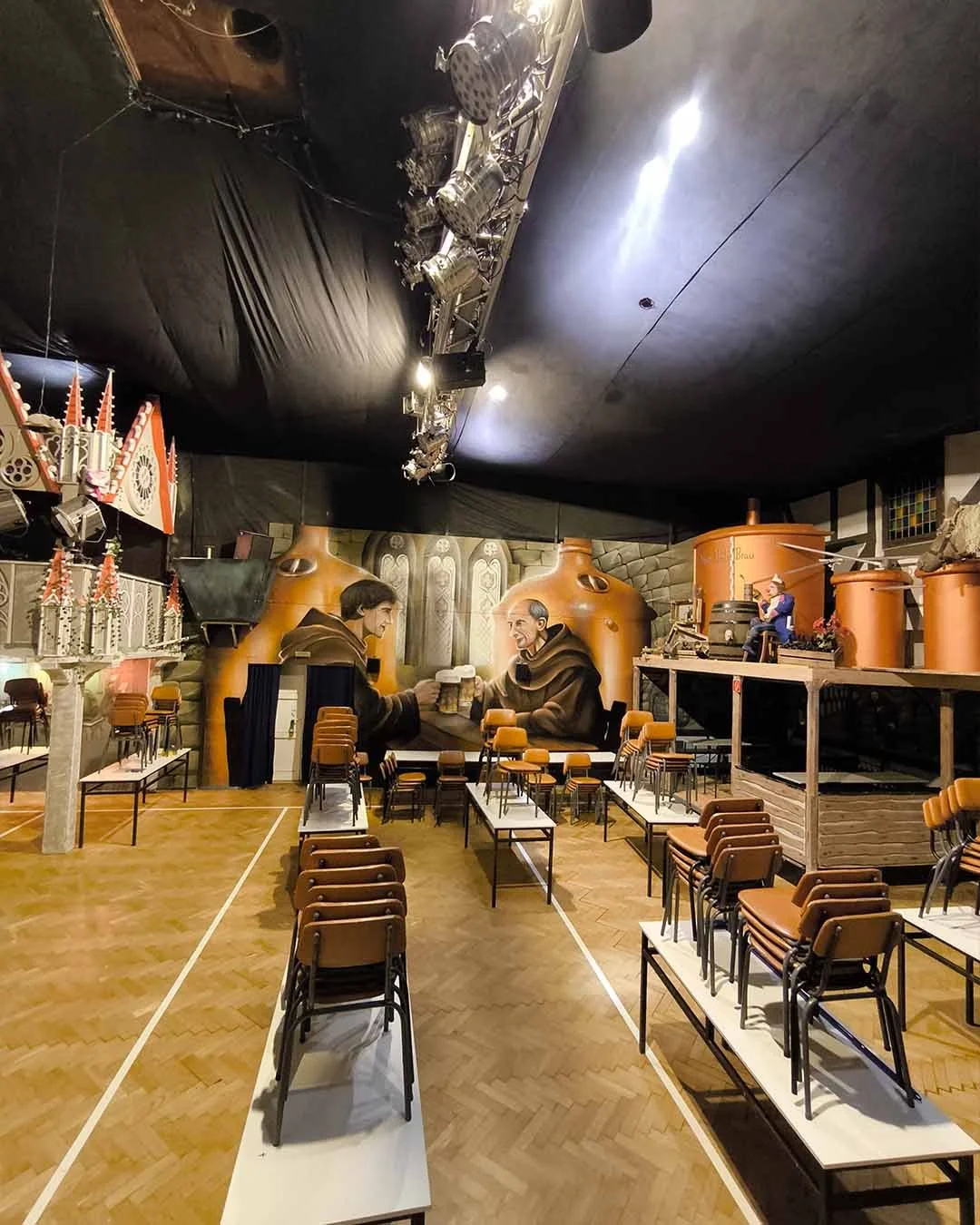 Innenansicht eines Raums, wahrscheinlich eines Gemeinderaums oder Veranstaltungssaals, mit einem großen Wandgemälde eines Mönchs, der Bier trinkt, in der Mitte der Wand. Der Raum ist mit Stühlen und Tischen ausgestattet, die für eine Veranstaltung vo