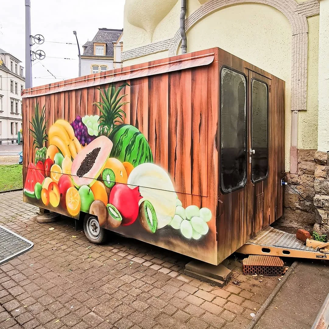 Kleinbus mit Fassade bemalt mit verschiedenen Früchten, wie Ananas, Wassermelone, Papaya, Kiwi, Orangen, Äpfel, Weintrauben und Honigmelone, vor einem Gebäude.