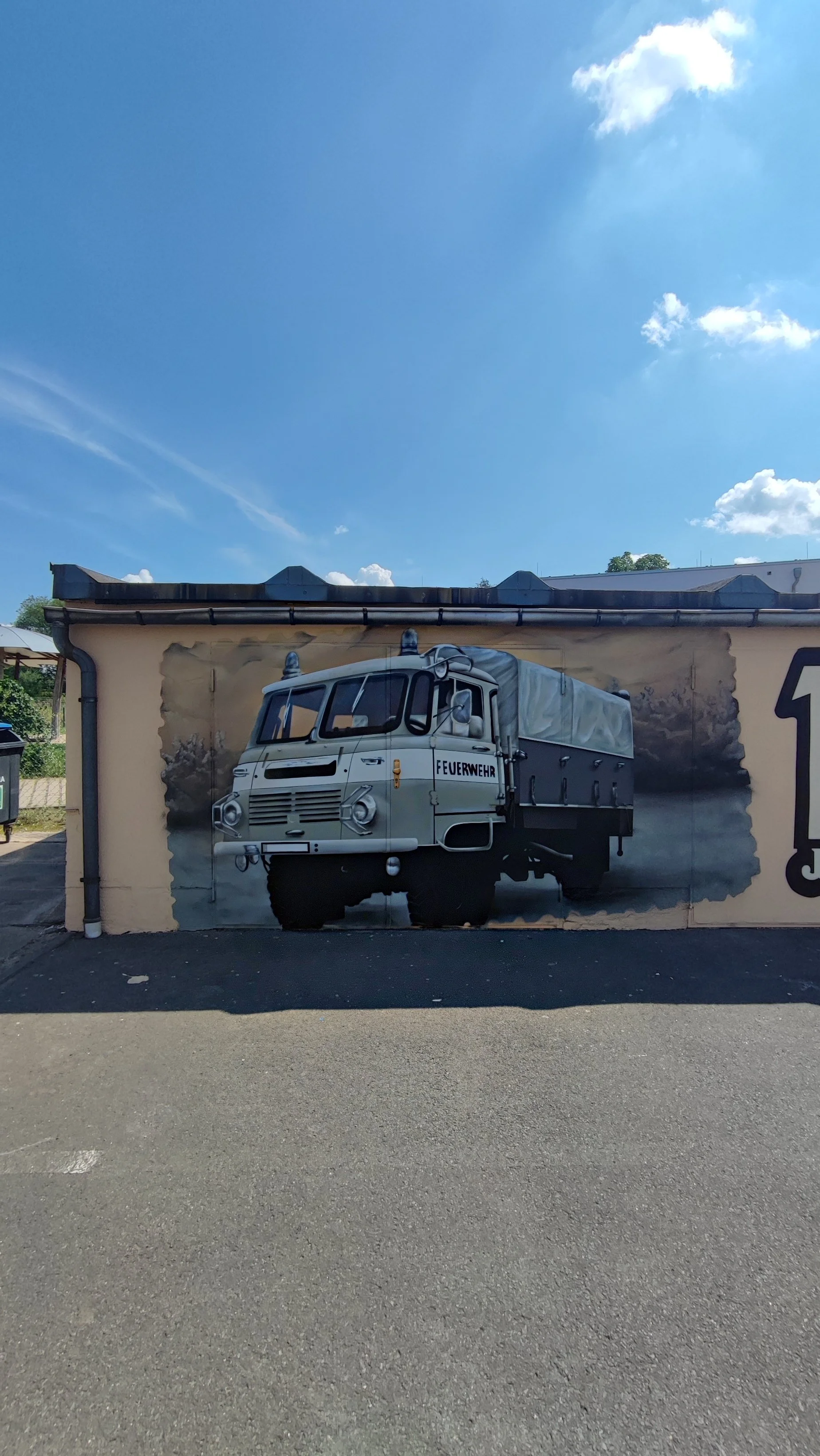 Wandbild eines alten Feuerwehrfahrzeugs mit dem Wort 'Feuerwehr' auf einer Hauswand. Im Hintergrund blauer Himmel mit Wolken.