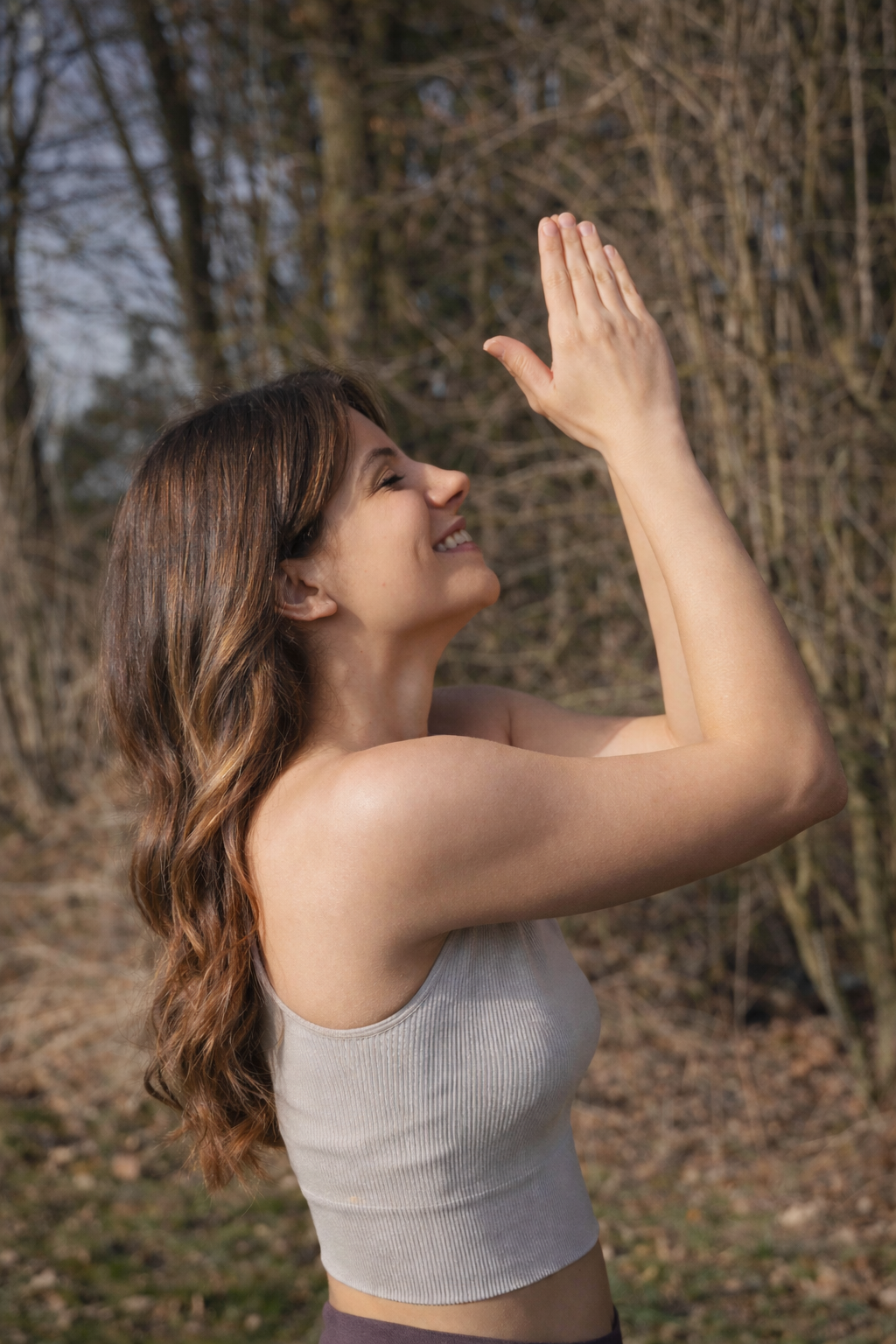 Eine lachende Frau mit braunen, welligen Haaren macht eine Yoga- oder Meditationsgeste im Freien vor Bäumen.
