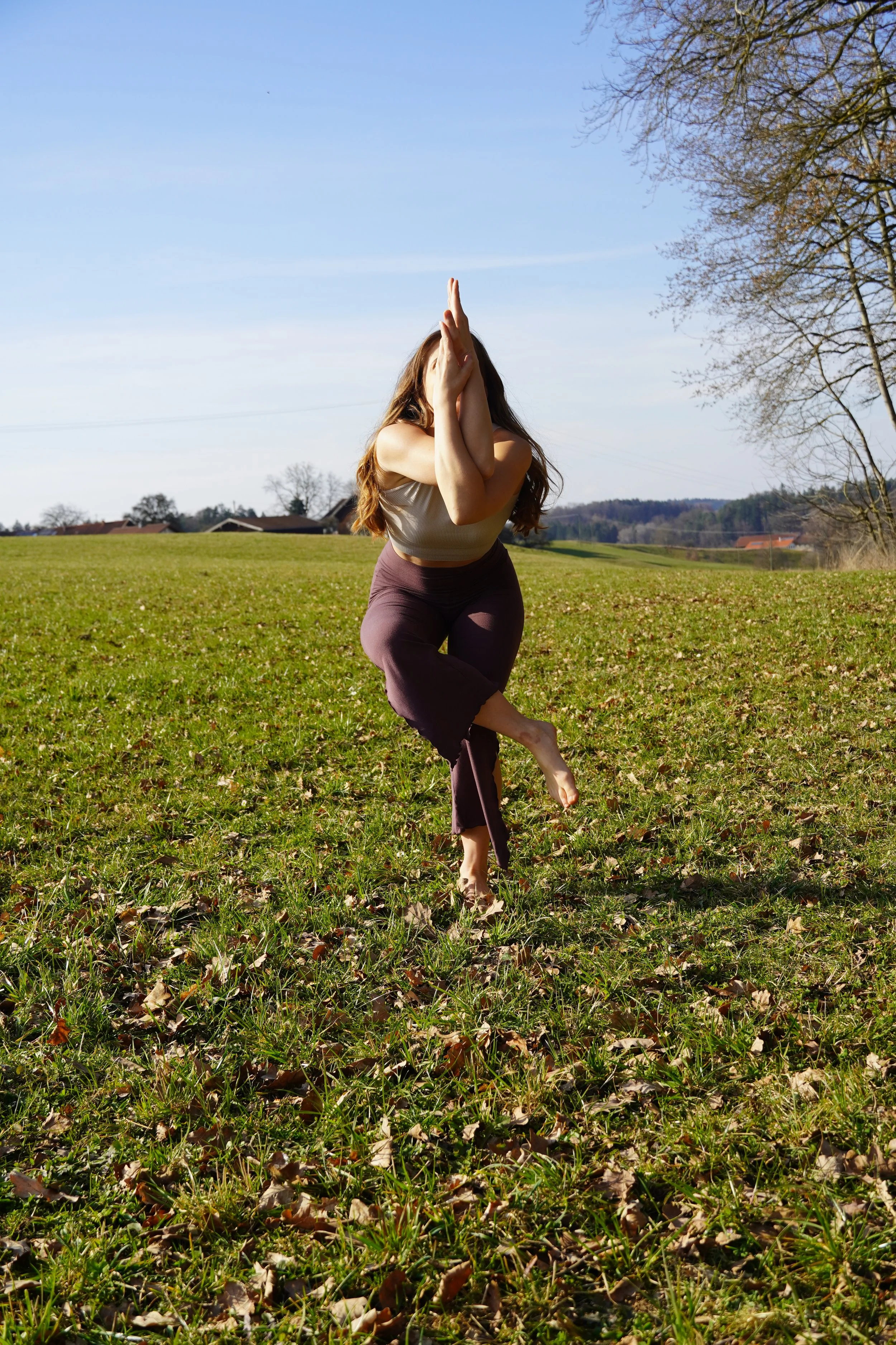 Frau, die in einer offenen Wiese Yoga macht, bei Sonnenlicht, im Freien, mit Bäumen und Häusern im Hintergrund.