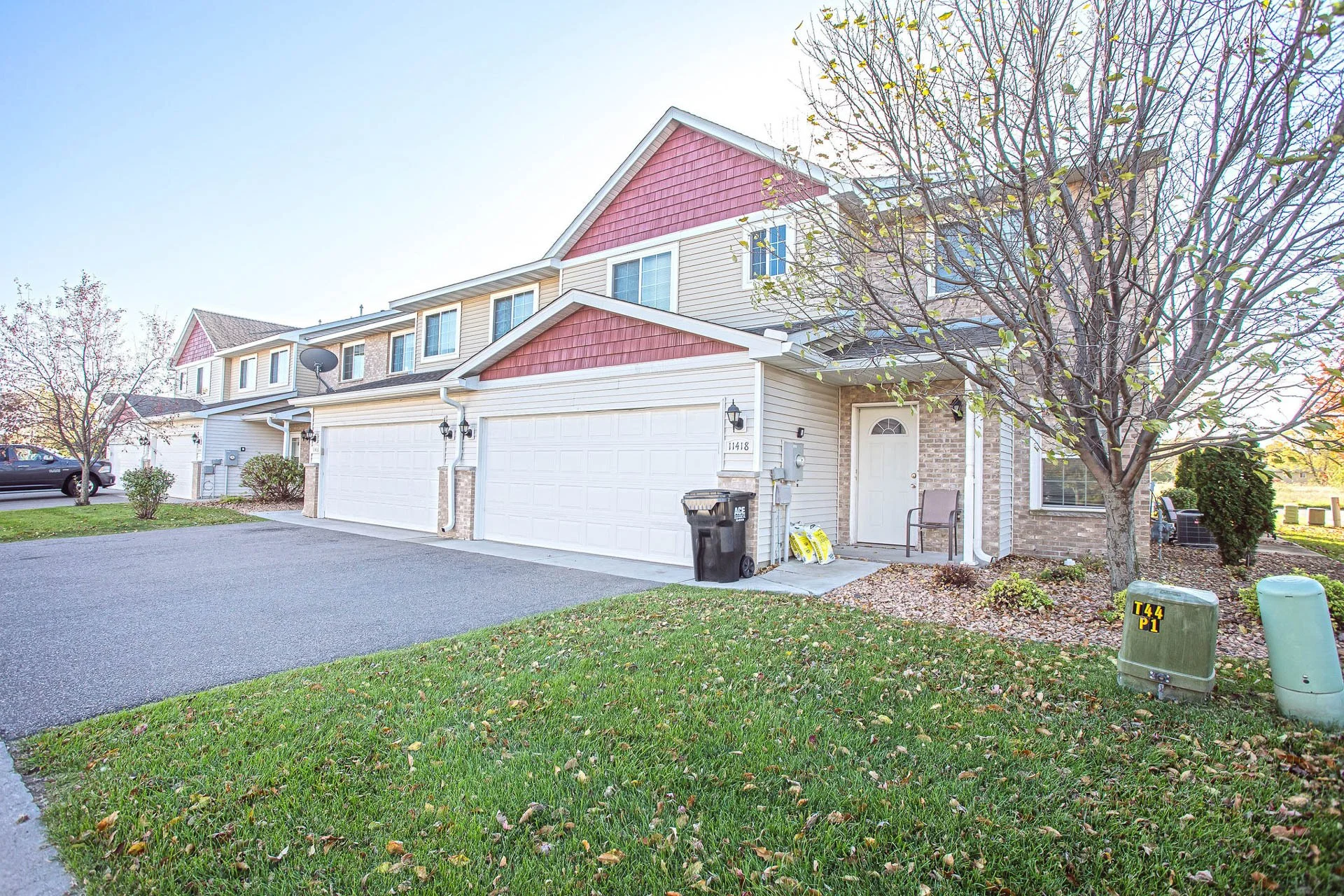 A two-story suburban house with a beige exterior, white garage doors, and pink decorative shingles on the upper part of the gables. There is a tree with yellowing leaves in front, a small porch with a chair, and a well-maintained lawn with fallen lea