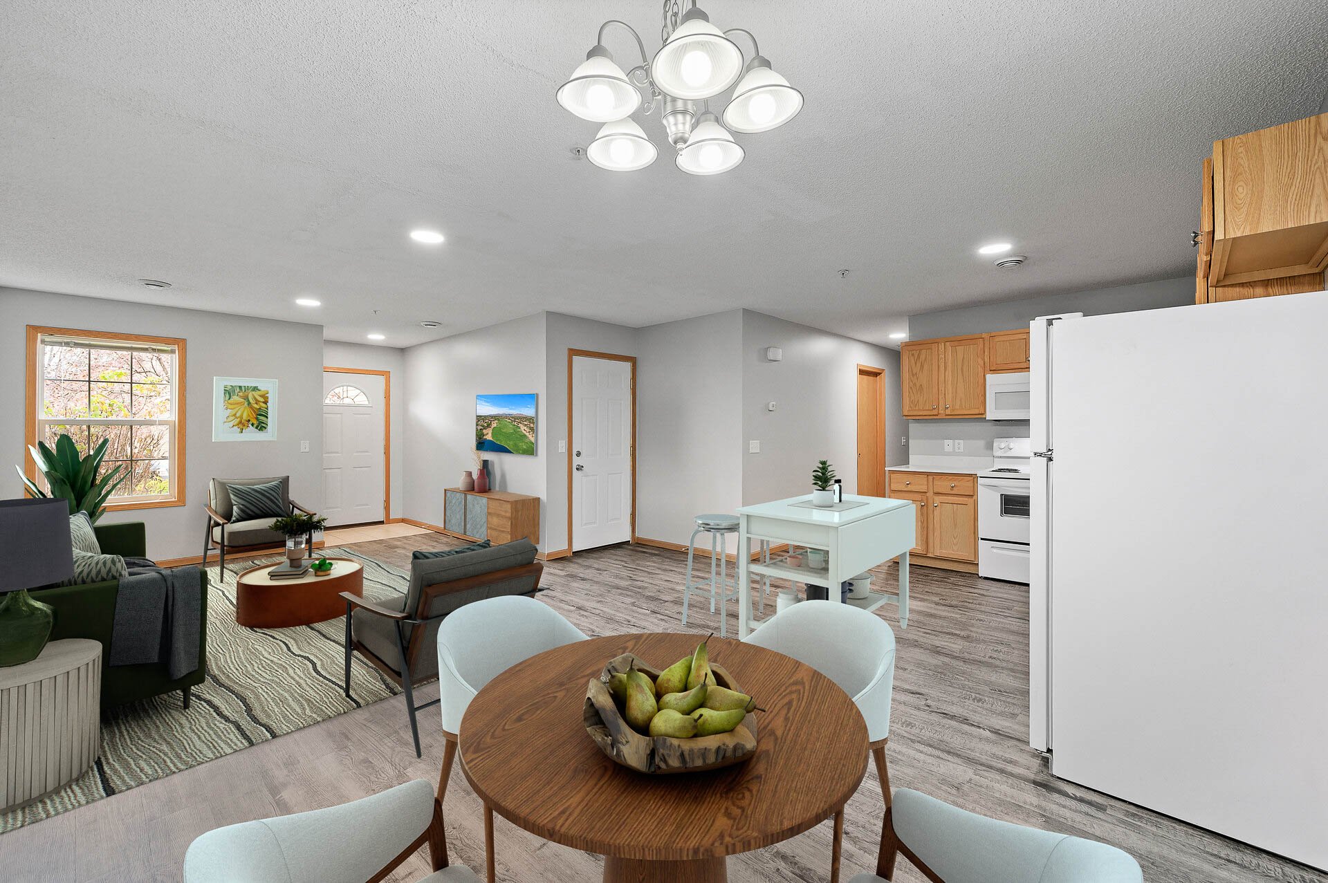 Open-concept living room and kitchen area with hardwood flooring, a round dining table with pears, white chairs, a kitchen with wooden cabinets, white appliances, and a small white table