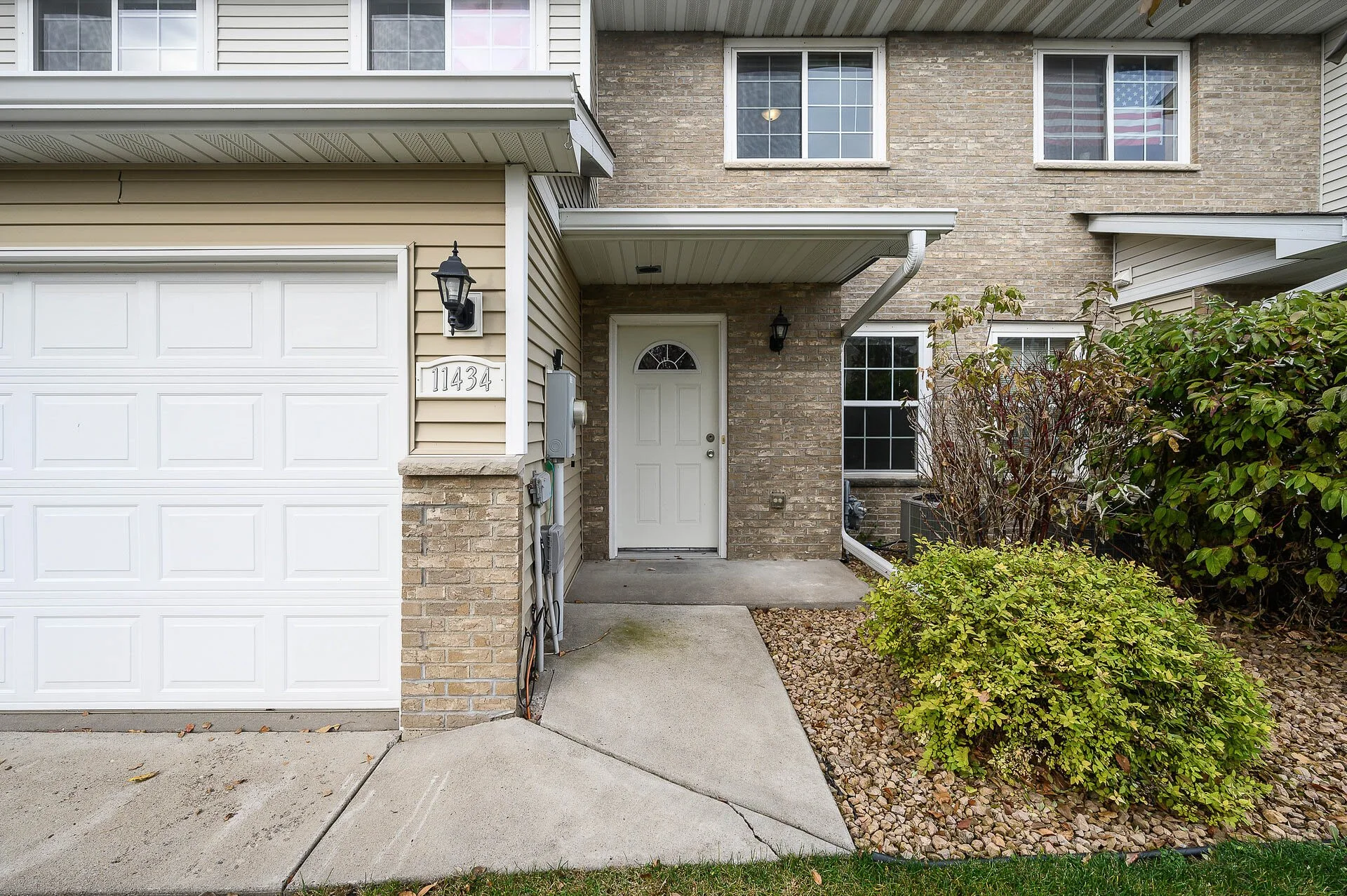Front entrance of a residential building with a white door, brick and siding exterior, and a small pathway leading to the door. There is a bush and gravel landscaping on the right side.