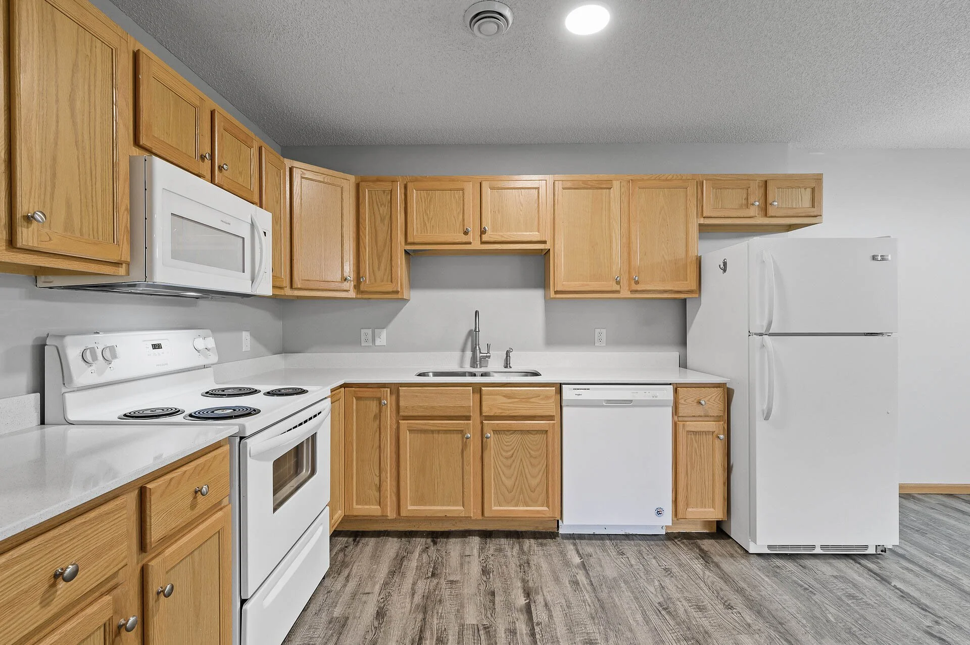 Kitchen with wooden cabinets, white appliances including microwave, stove, dishwasher, and refrigerator, gray walls, and wood-look flooring.