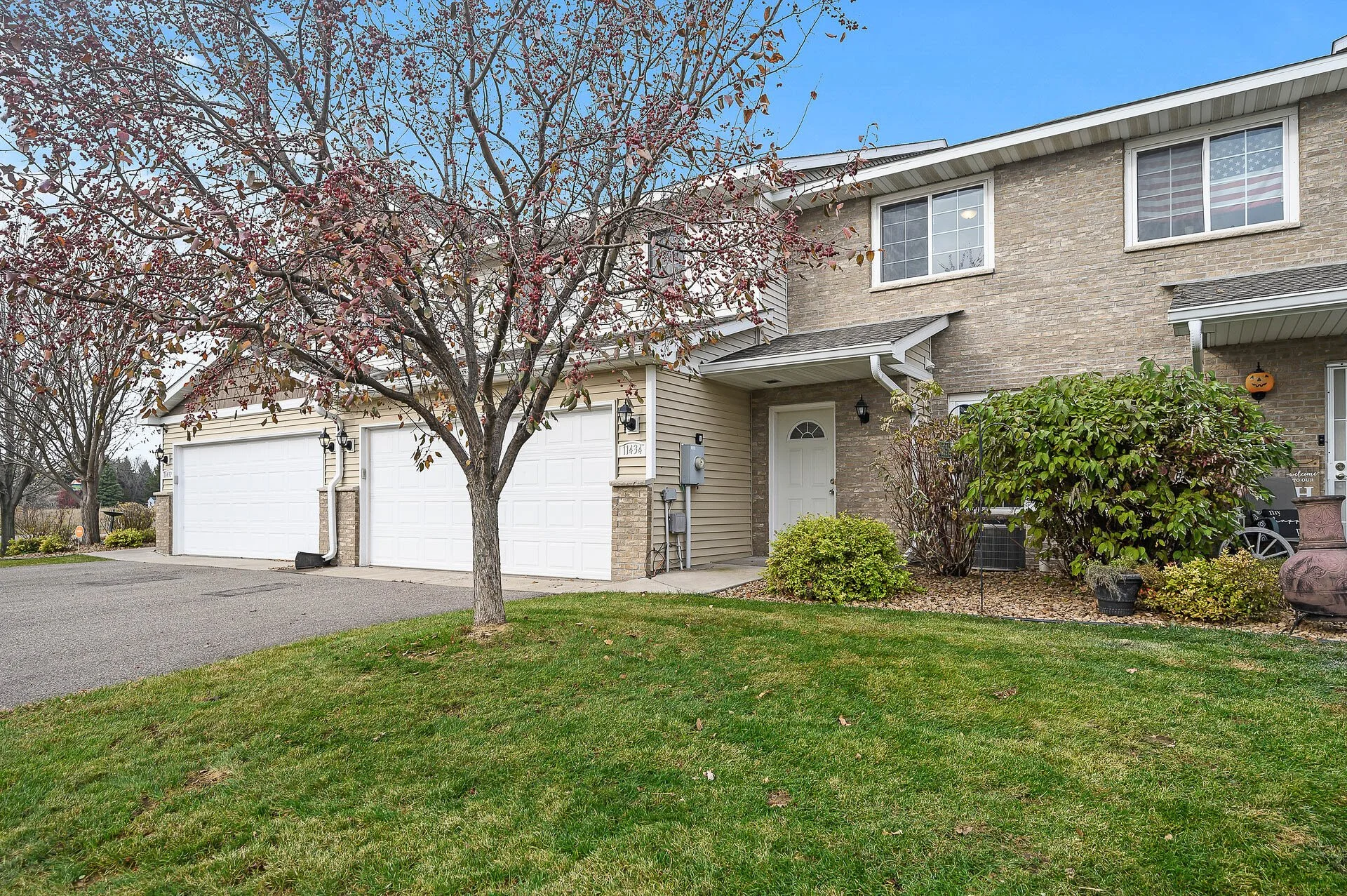 Front view of a two-story house with a garage, a tree with red leaves, green lawn, and shrubs under a blue sky.
