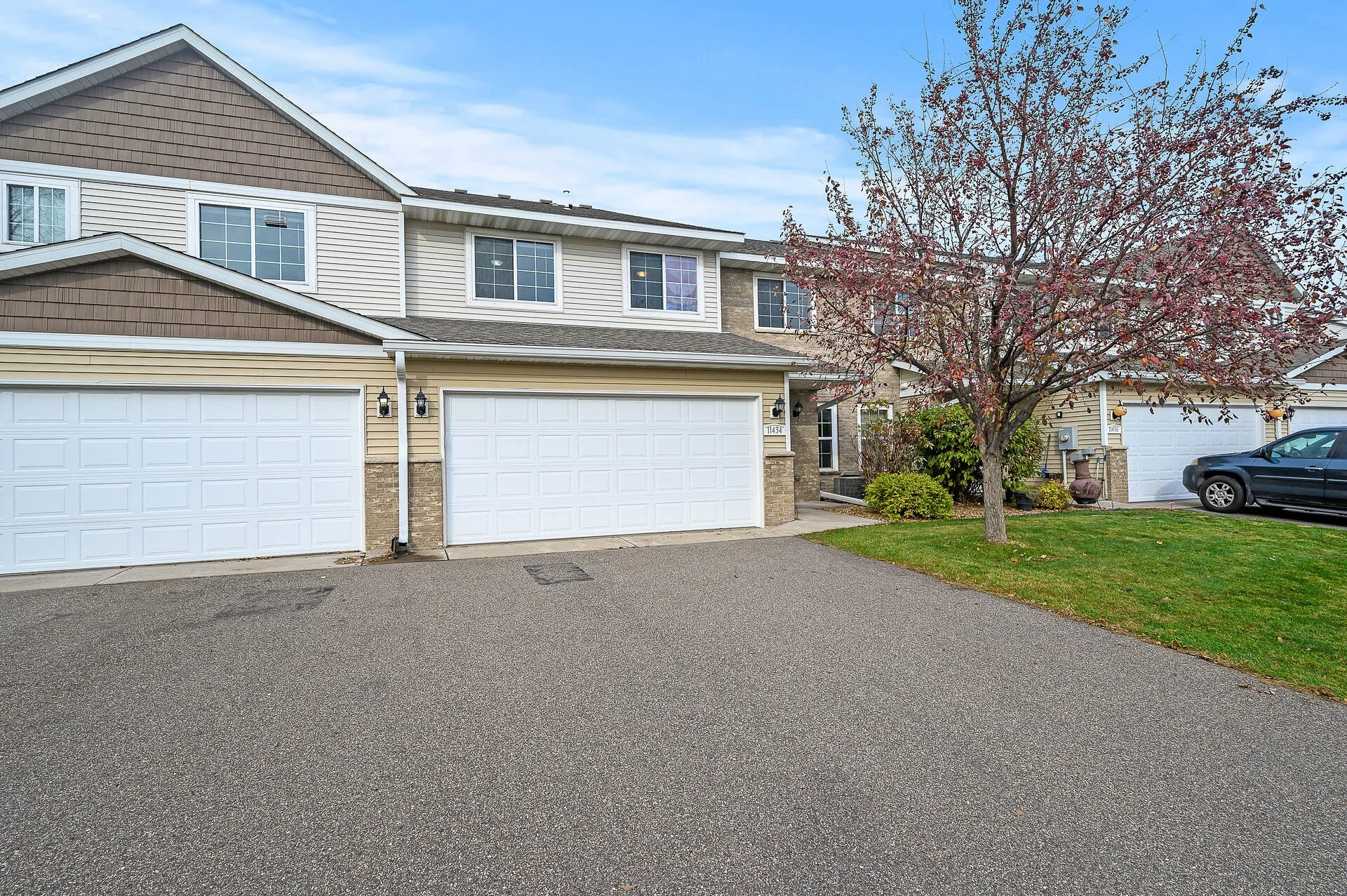 A residential apartment complex with a multi-car garage, a tree with fall foliage, and a parked black car in the driveway.