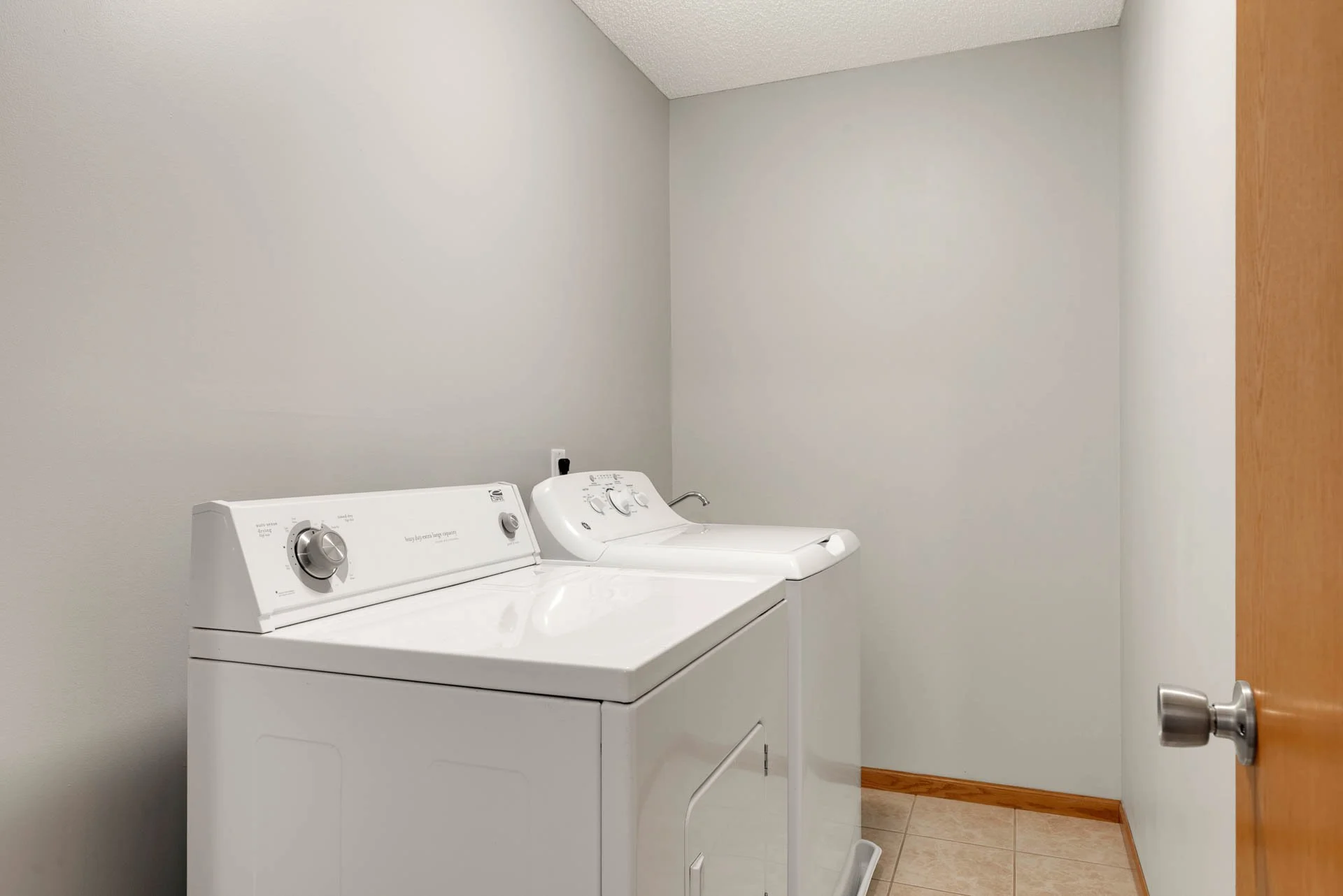 A laundry room with a washing machine and a dryer beside it, both white, against a plain gray wall, with beige tiled flooring and a wooden door on the right.