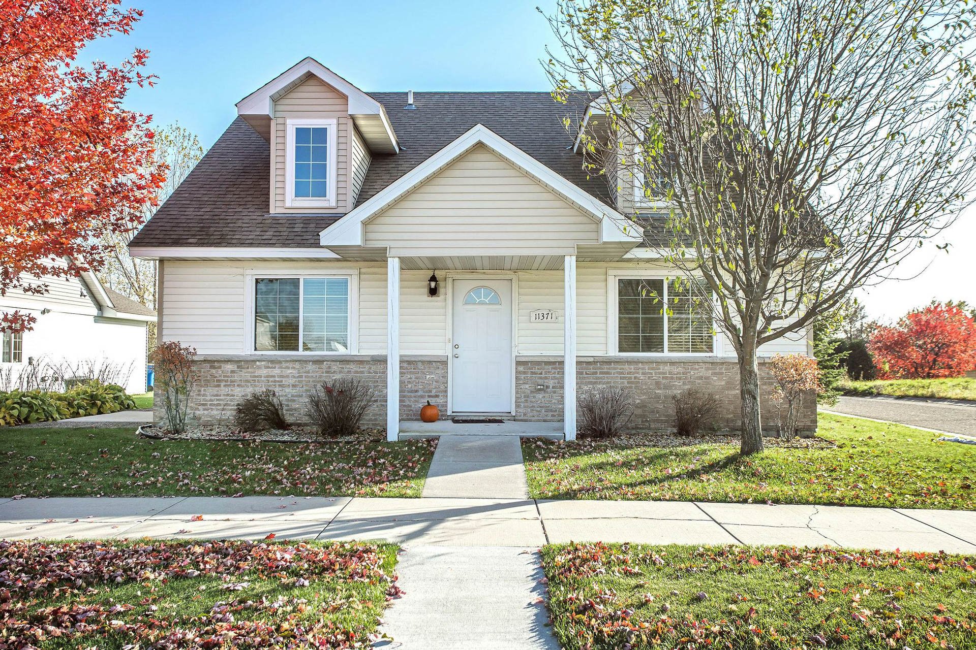 Front view of a house with white siding, brick foundation, and a gray shingle roof, surrounded by trees with fall foliage, and a well-maintained lawn.