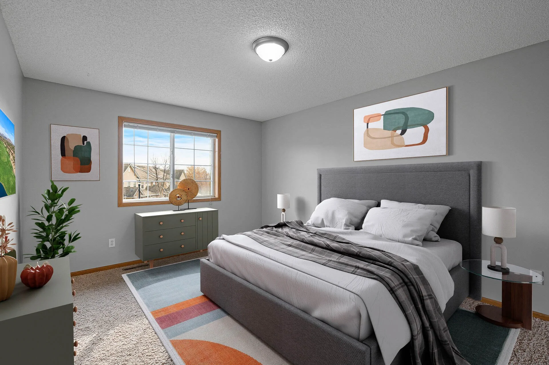 A bedroom with a gray bed, white bedding, and a plaid throw, sunset view through a window, abstract art above the bed, green dresser, side table with lamp, colorful area rug, and a plant in the corner.