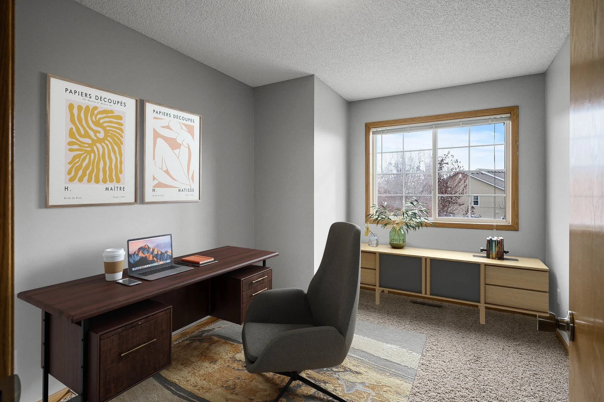 Home office with a wooden desk, grey chair, framed art prints on the wall, a window with a view of a neighborhood, and a wooden storage bench with a plant and books.