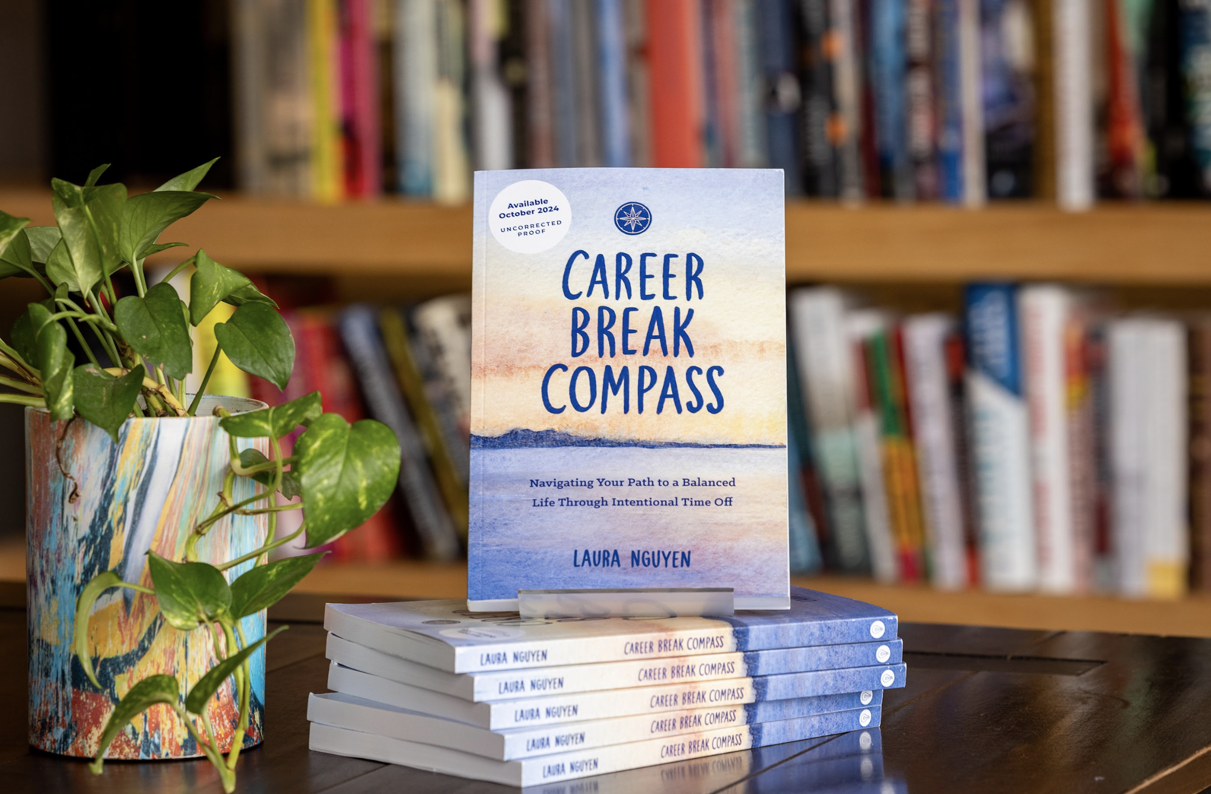 Stack of books titled "Career Break Compass" by Laura Nguyen with a colorful landscape cover and a small potted plant on a wooden surface, in front of a background of a bookshelf.