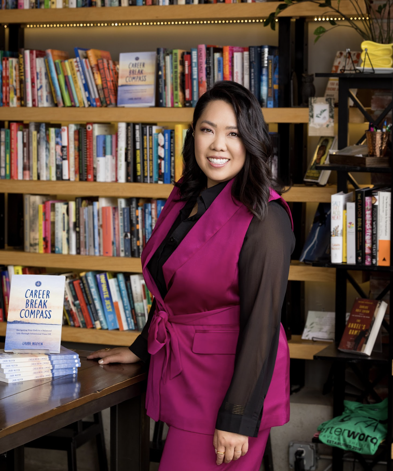 A woman with black, wavy hair dressed in a hot pink blazer and matching pants, standing next to a table with a book titled 'Career Break Compass' in a bookstore or library with wooden shelves full of colorful books in the background.