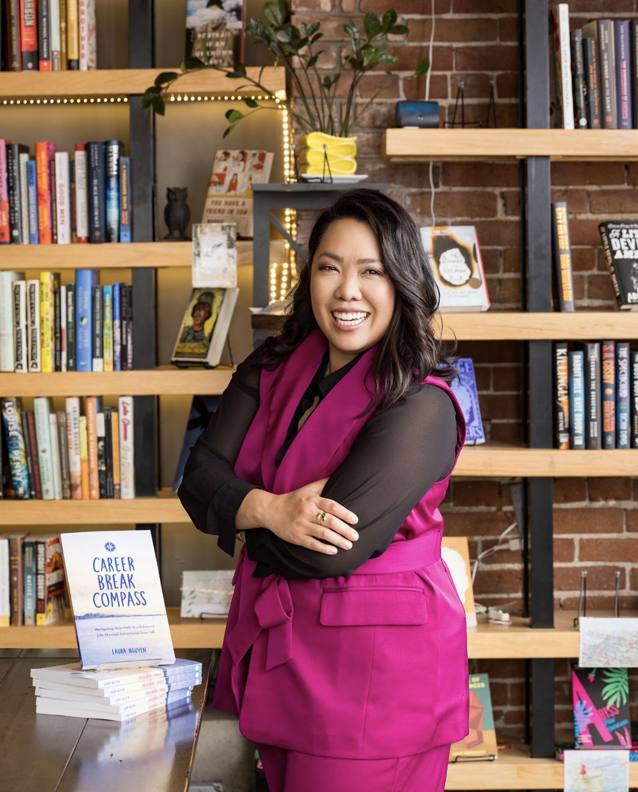 A woman with long dark hair wearing a black top and a bright pink vest standing in front of bookshelves, smiling with one eye winking, in a bookstore or library setting.