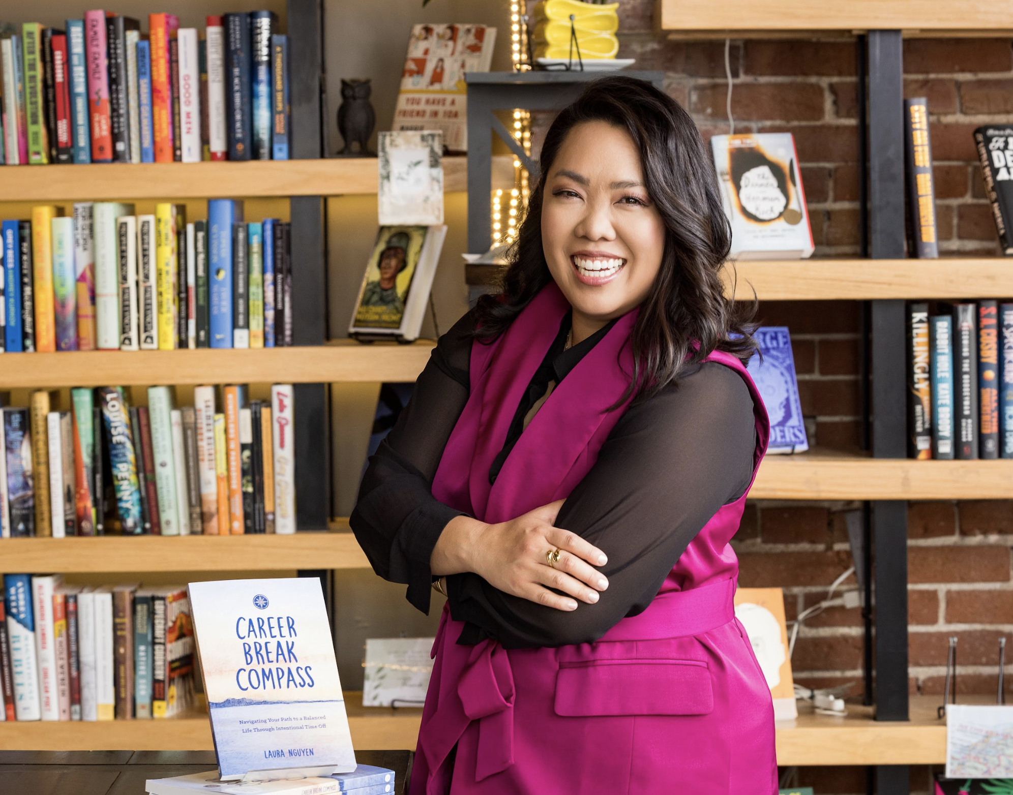A smiling woman with dark hair wearing a black top and pink vest, standing in front of a bookshelf with colorful books, and a table with a book titled "Career Break Compass" by Laura Nguyen.
