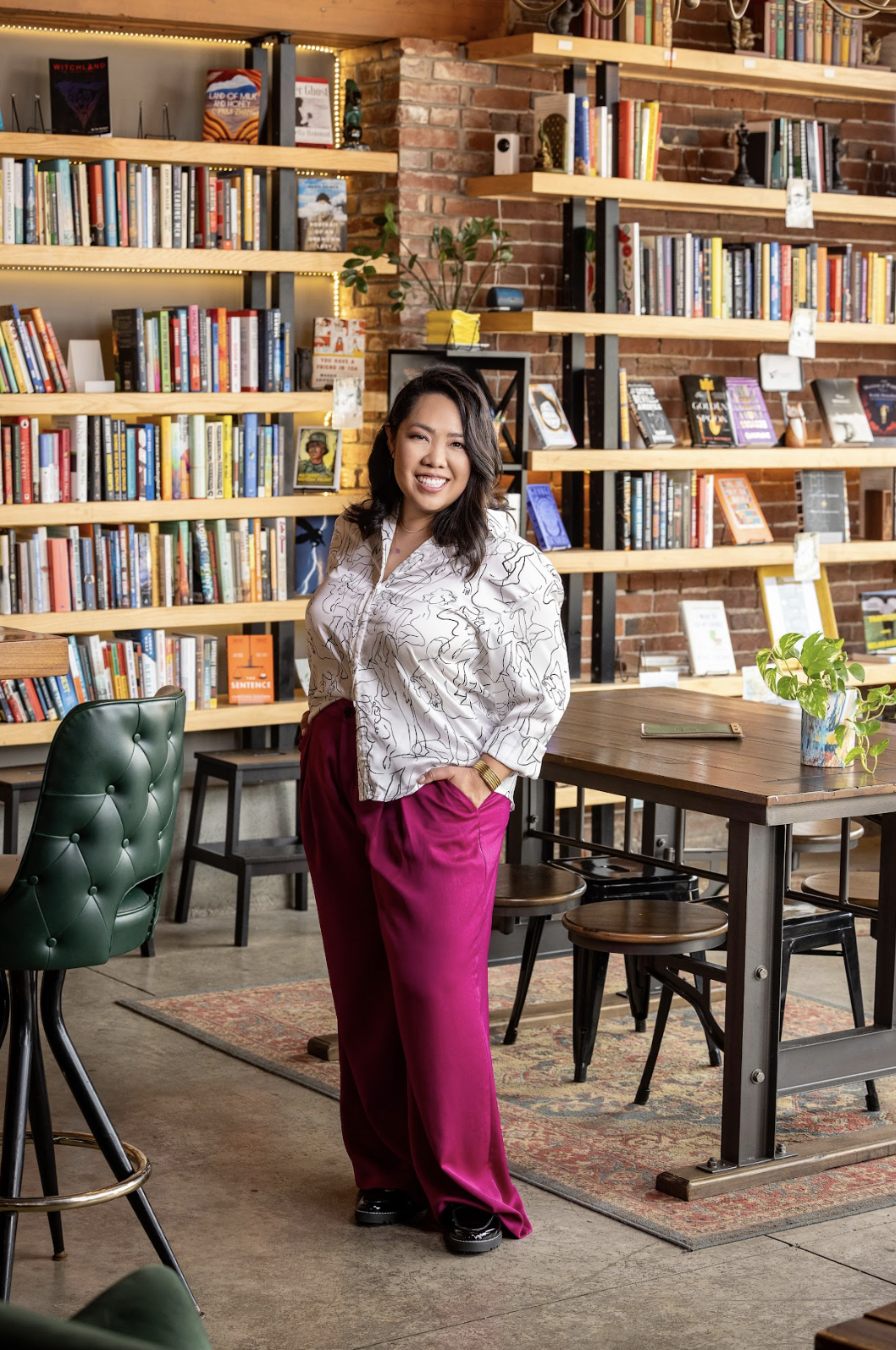 A smiling woman standing in a cozy, well-lit bookstore or cafe with wooden shelves filled with books behind her. She has shoulder-length dark hair, is wearing a patterned white blouse and bright pink pants, and is standing next to a wooden table with plants.