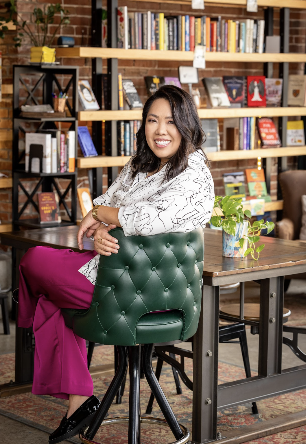 A woman with dark hair and a silver bracelet sitting on a green tufted chair in a bookstore or library, smiling and looking at the camera. Behind her are wooden shelves filled with books, and there is a potted plant on the table to her right.