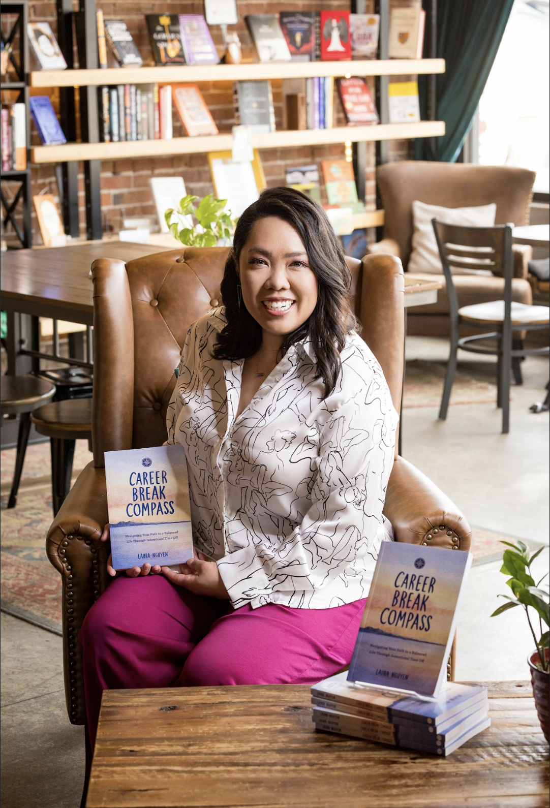 A woman sitting on a leather armchair holding a book titled 'Career Break Compass,' smiling and looking at the camera. There are more copies of the book on a wooden table in front of her, in a cozy bookstore or cafe setting with bookshelves, chairs, and a brick wall in the background.