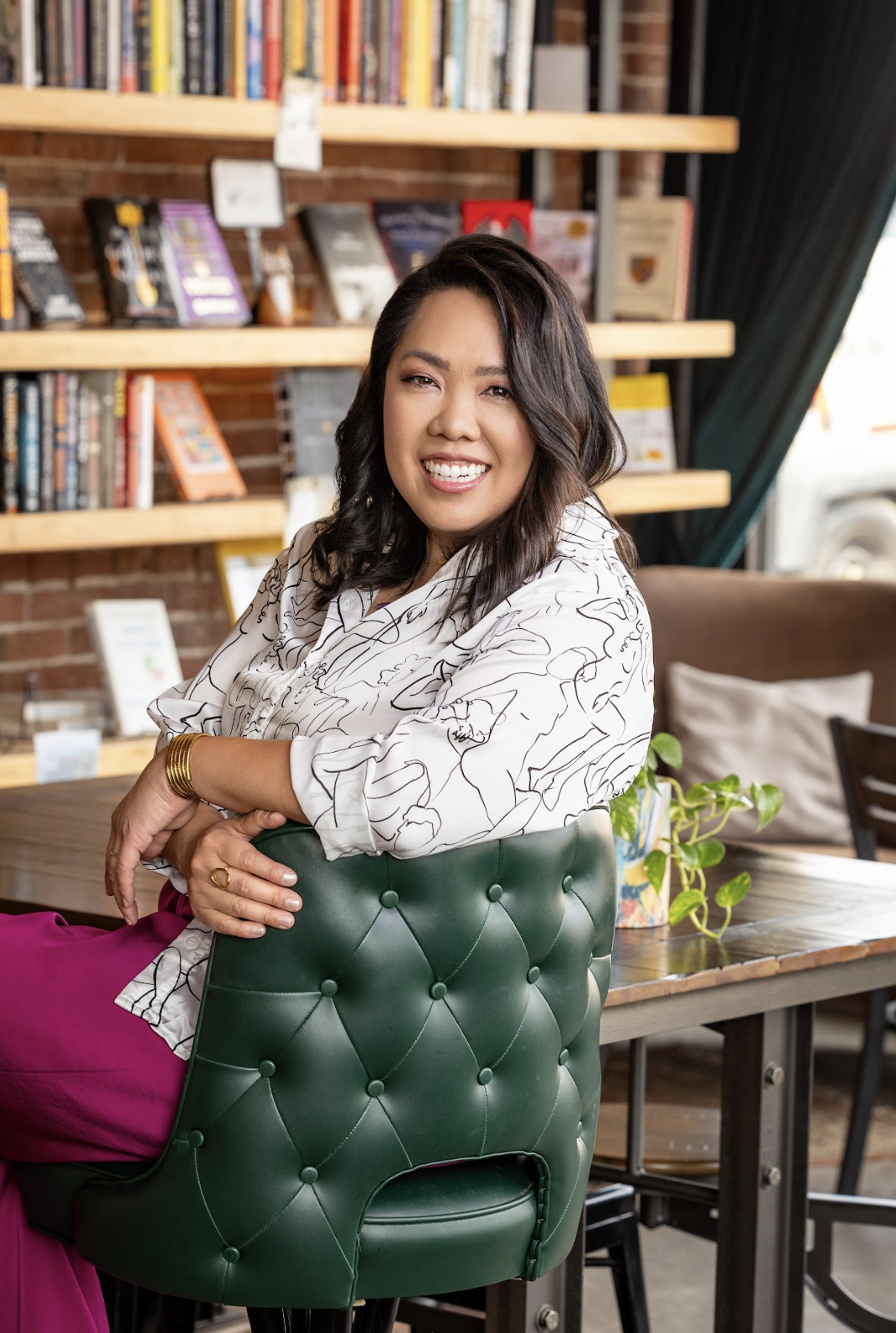 A woman with dark wavy hair smiling and sitting on a green tufted chair in a cozy bookstore or cafe, with shelves of books and a brick wall in the background.