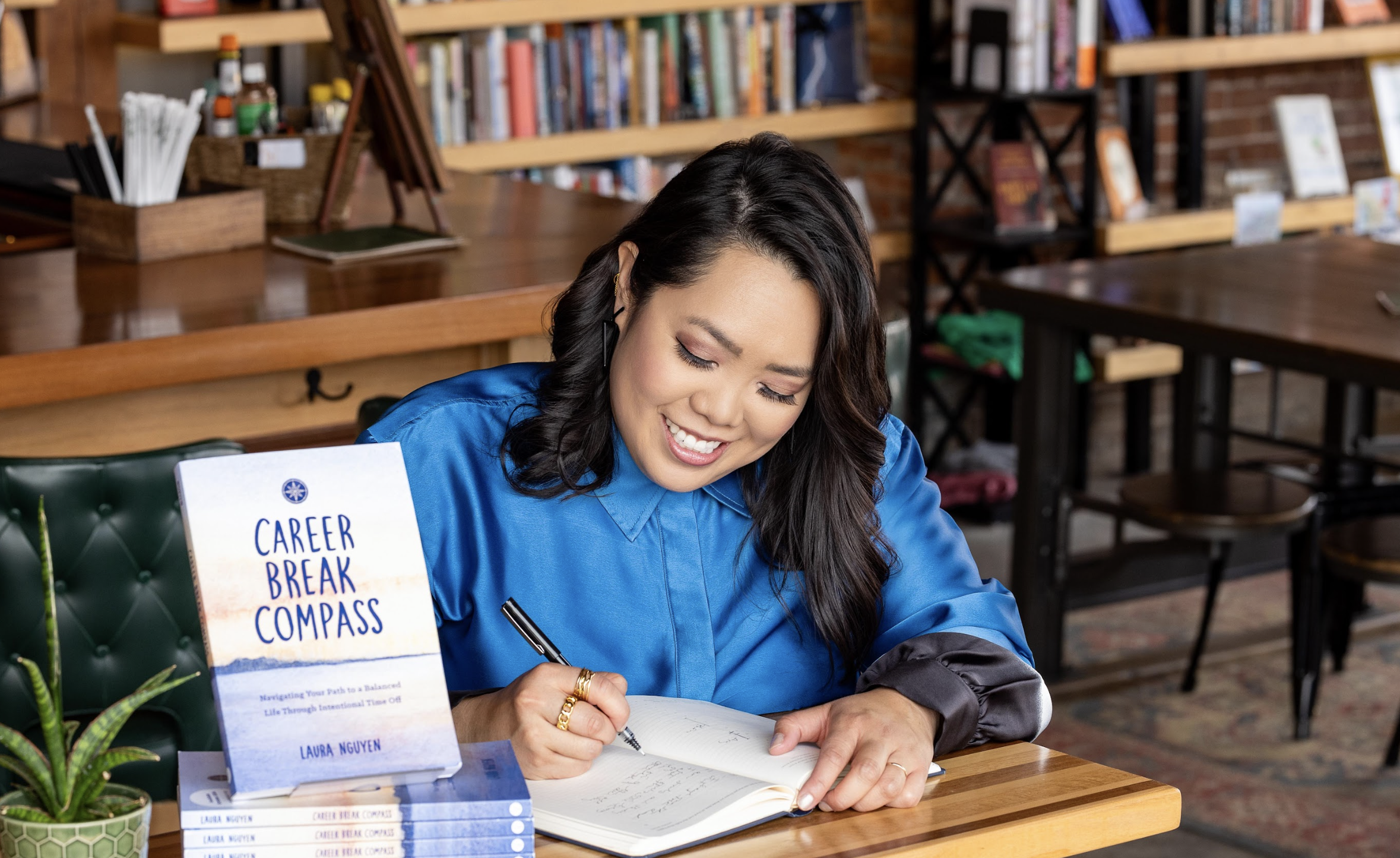 A woman in a blue shirt is writing in a notebook at a wooden desk, smiling. A book titled 'Career Break Compass' by Laura Nguyen is on the desk.
