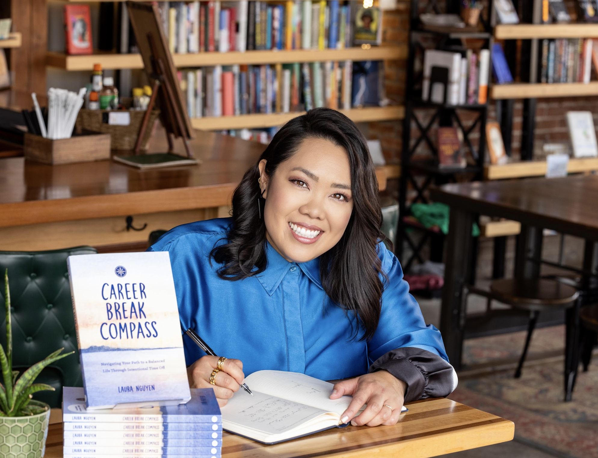 Woman sitting at a table in a bookstore, smiling, with a book titled 'Career Break Compass' by Laura Nguyen, and an open notebook in front of her.