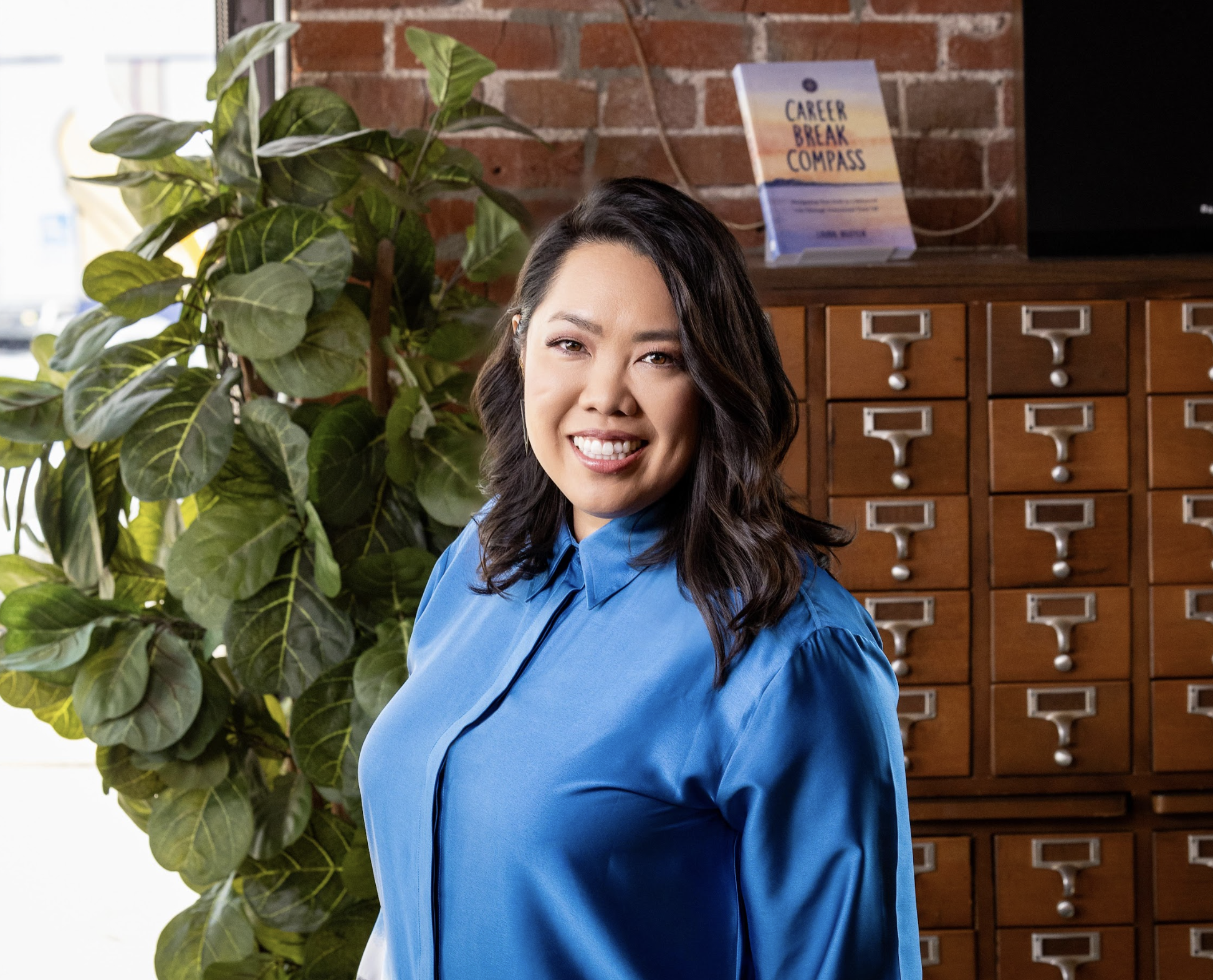 A woman with shoulder-length dark hair smiling and wearing a blue shirt standing indoors near a large green plant and a wooden card catalog cabinet with small drawers, in front of a brick wall. A sign on top of the cabinet reads 'Career Break Compass'.