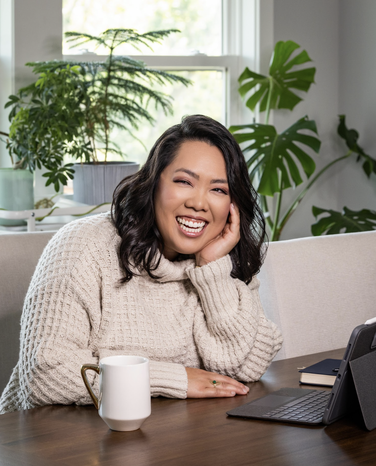 A woman with black hair smiling and winking at a laptop on a desk, surrounded by green houseplants, inside a bright room.
