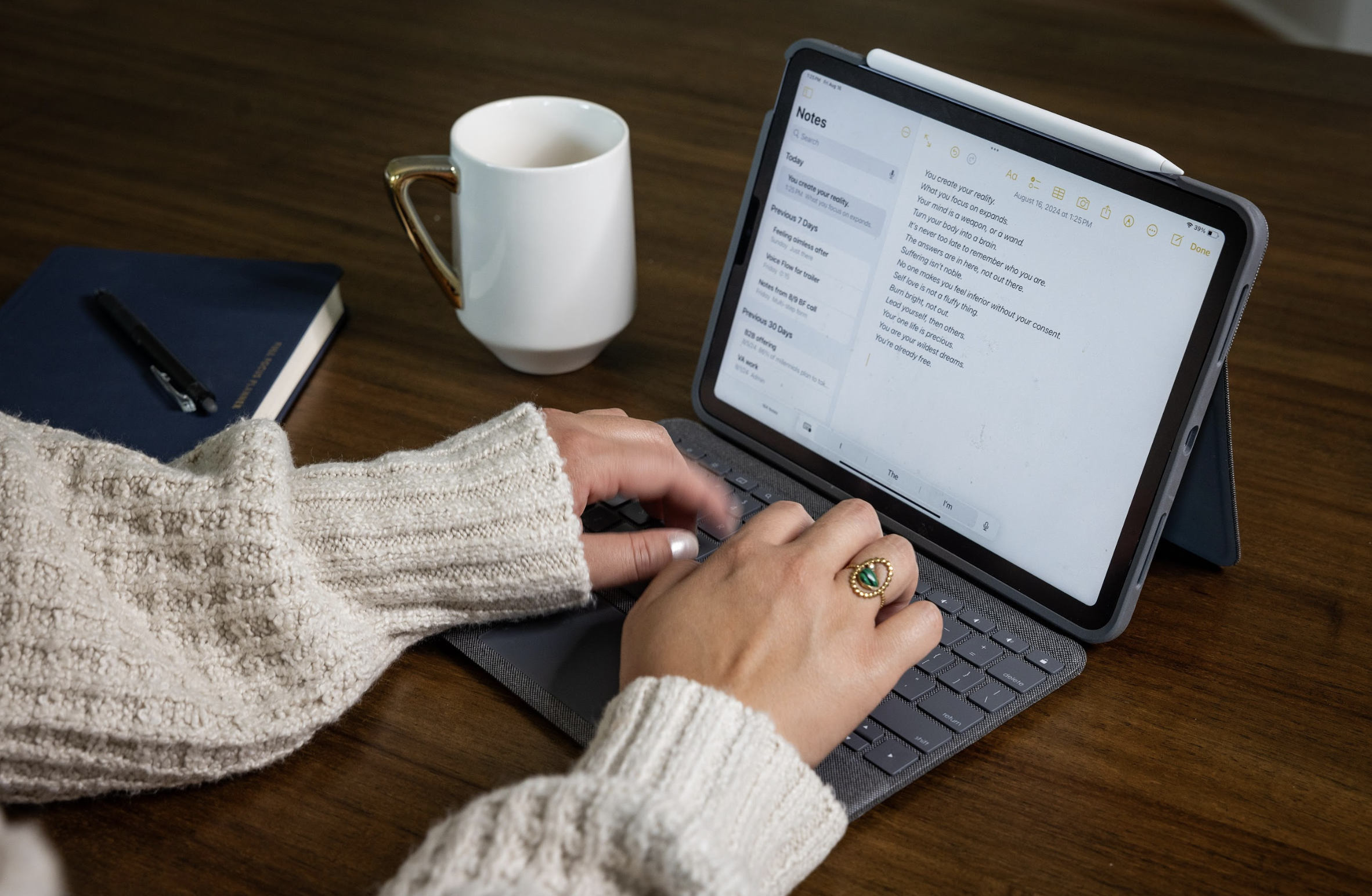 Person wearing a beige sweater typing on a tablet with a keyboard, a white mug, a closed blue notebook, and a pen on a wooden table.